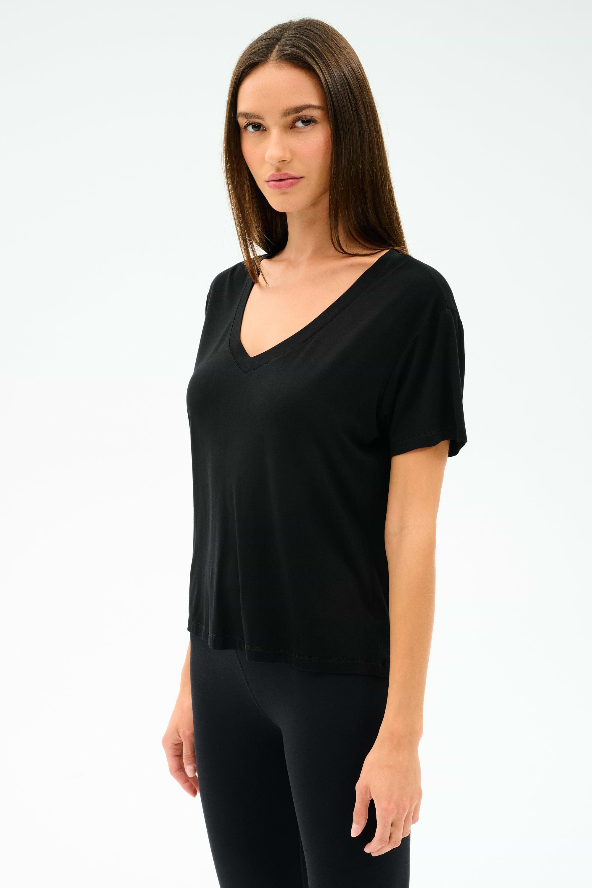 A woman with long straight hair wears the SPLITS59 Georgia Jersey V-Neck in black, styled with black leggings, standing against a plain light background.