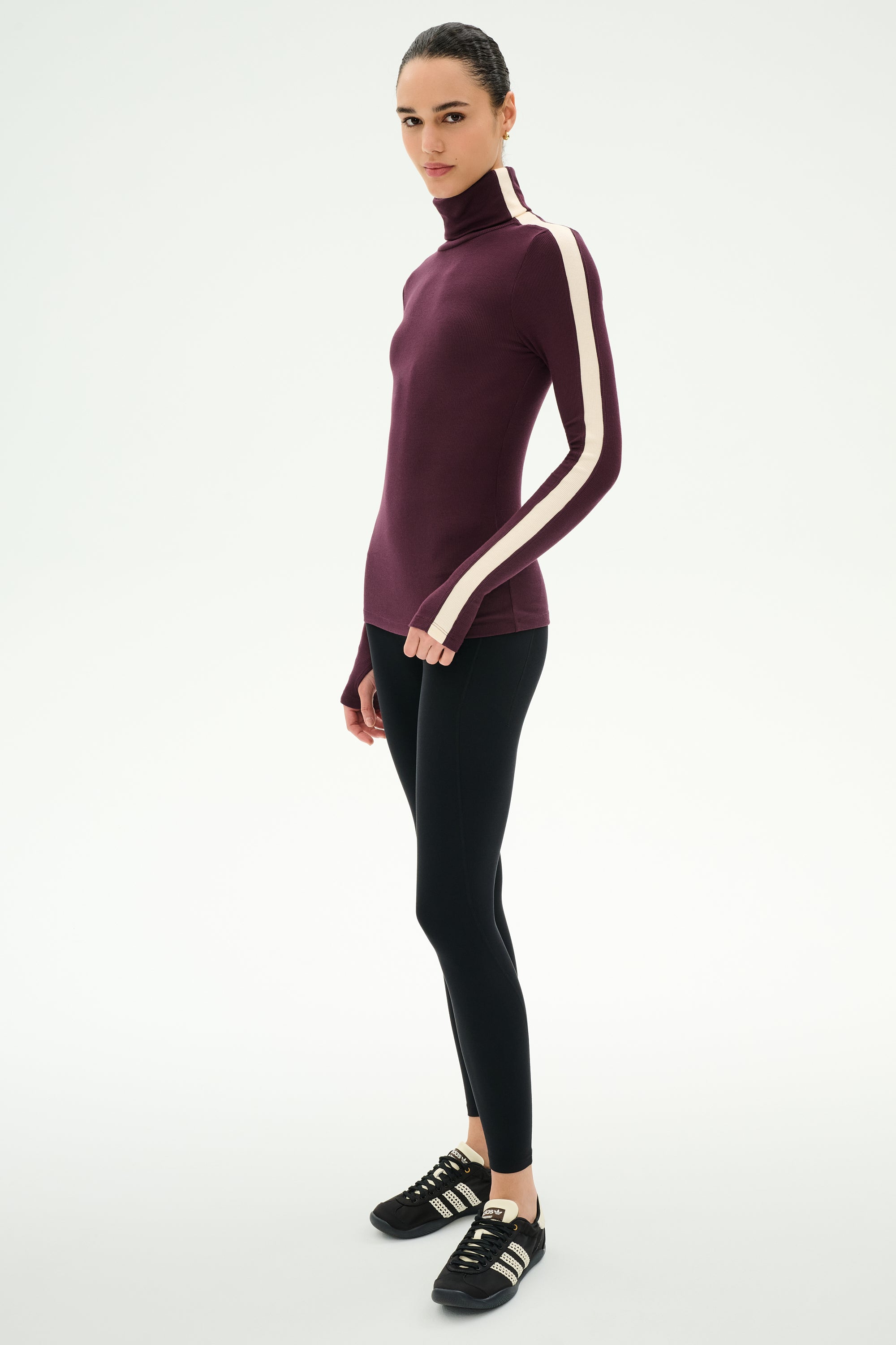 A person wears the SPLITS59 Jackson Rib Full Length Turtleneck in Dark Cherry/Creme, paired with black modal blend leggings featuring spandex for added stretch, and black sneakers, standing against a plain white background.