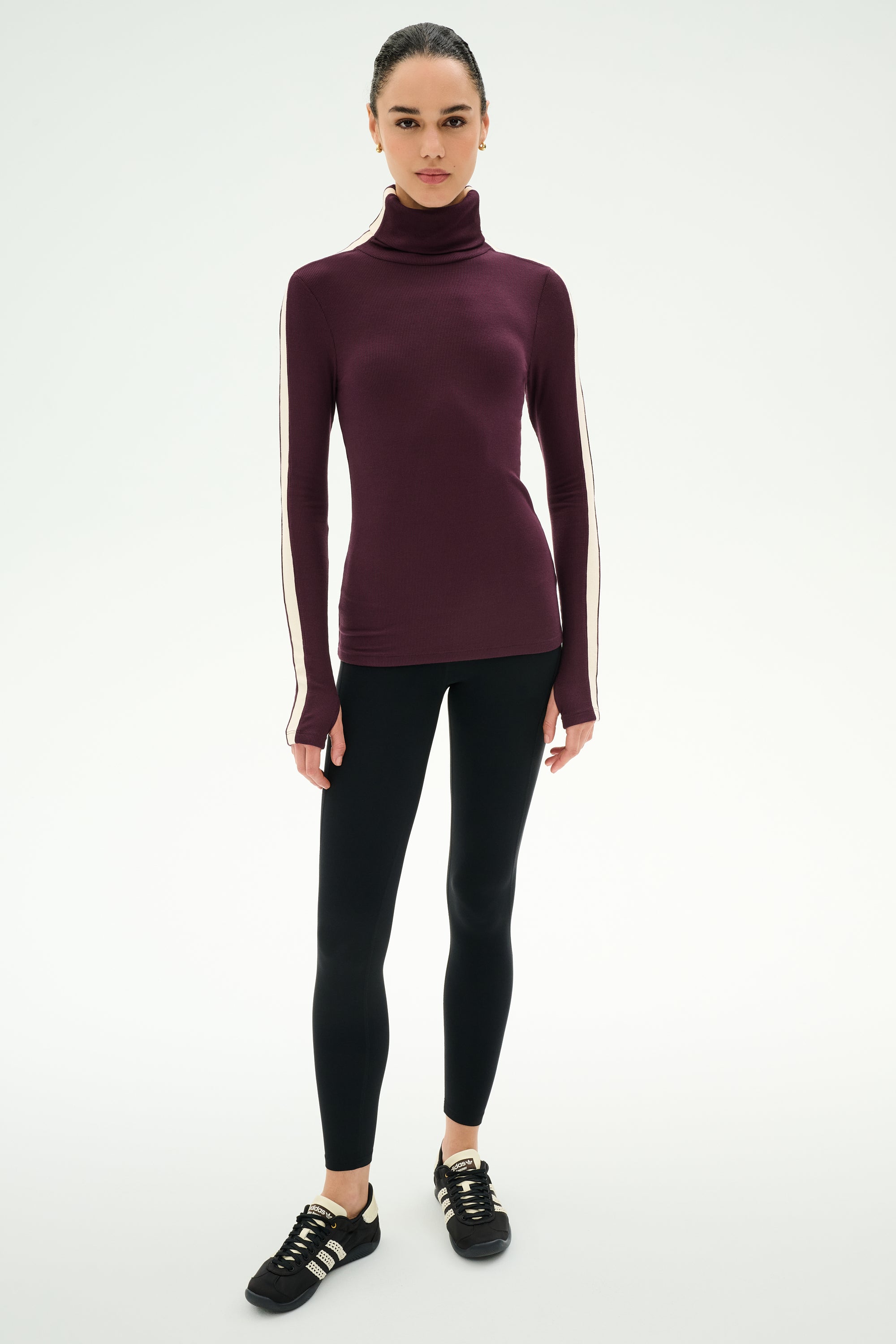A woman wears the SPLITS59 Jackson Rib Full Length Turtleneck in Dark Cherry/Creme—a maroon top with white striped sleeves, crafted from soft modal fabric—paired with black leggings and black sneakers, standing against a plain white background.