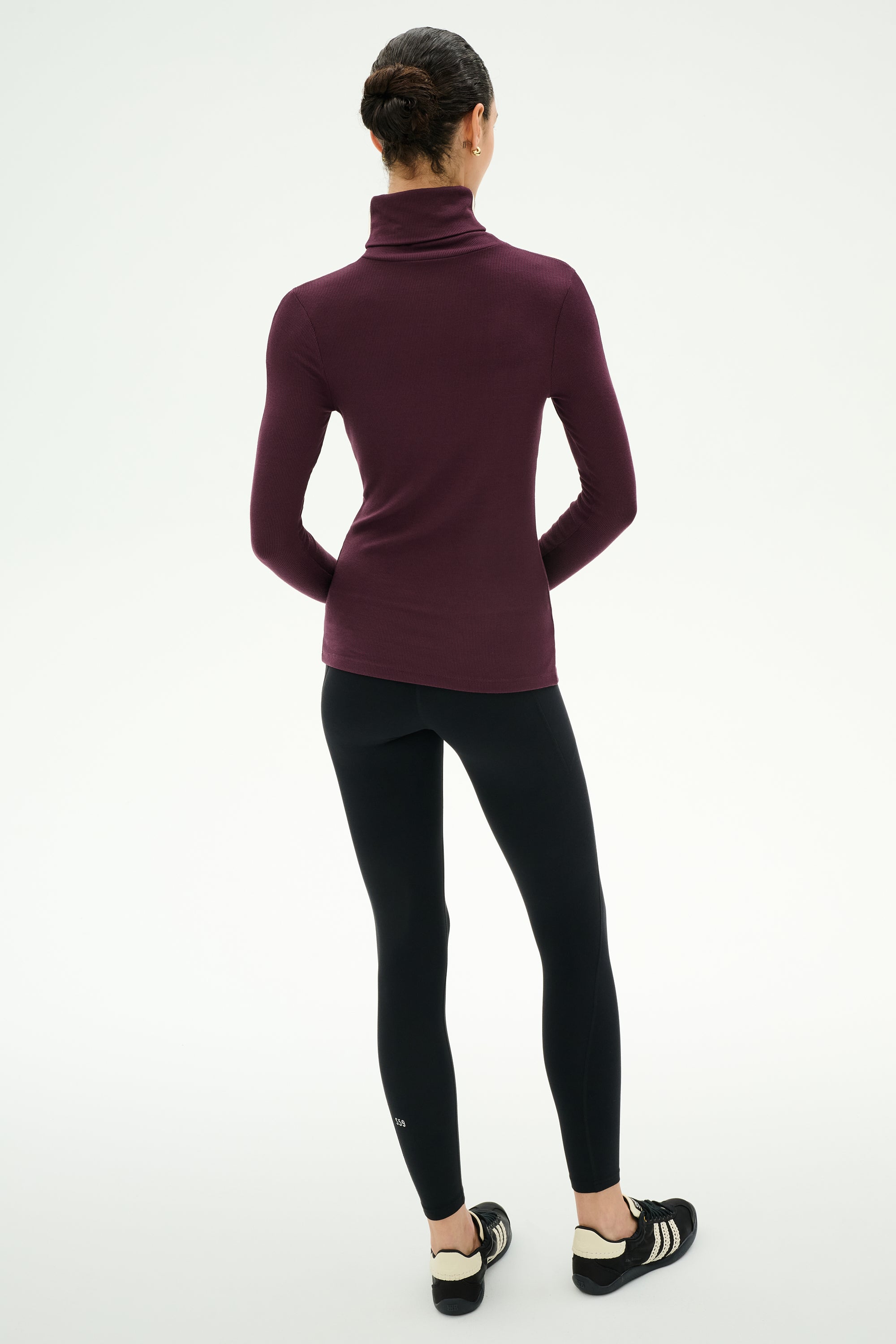 A person stands with their back to the camera, wearing the SPLITS59 Jackson Rib Full Length Turtleneck in Dark Cherry/Creme, paired with black leggings and black sneakers against a plain white background.