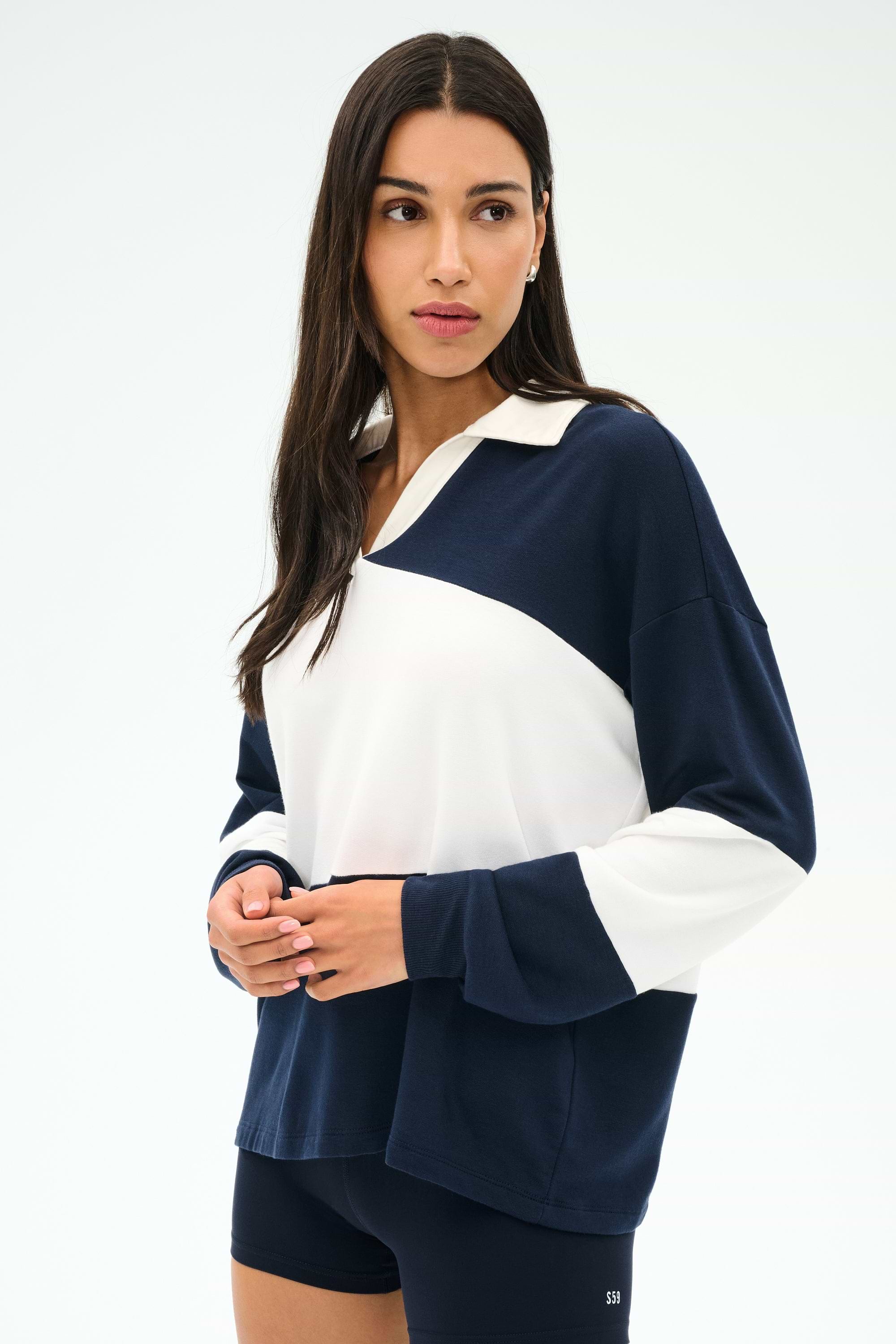 A woman with long dark hair wears the SPLITS59 Katie Fleece Rugby in Indigo/White and navy shorts, standing against a plain light background.