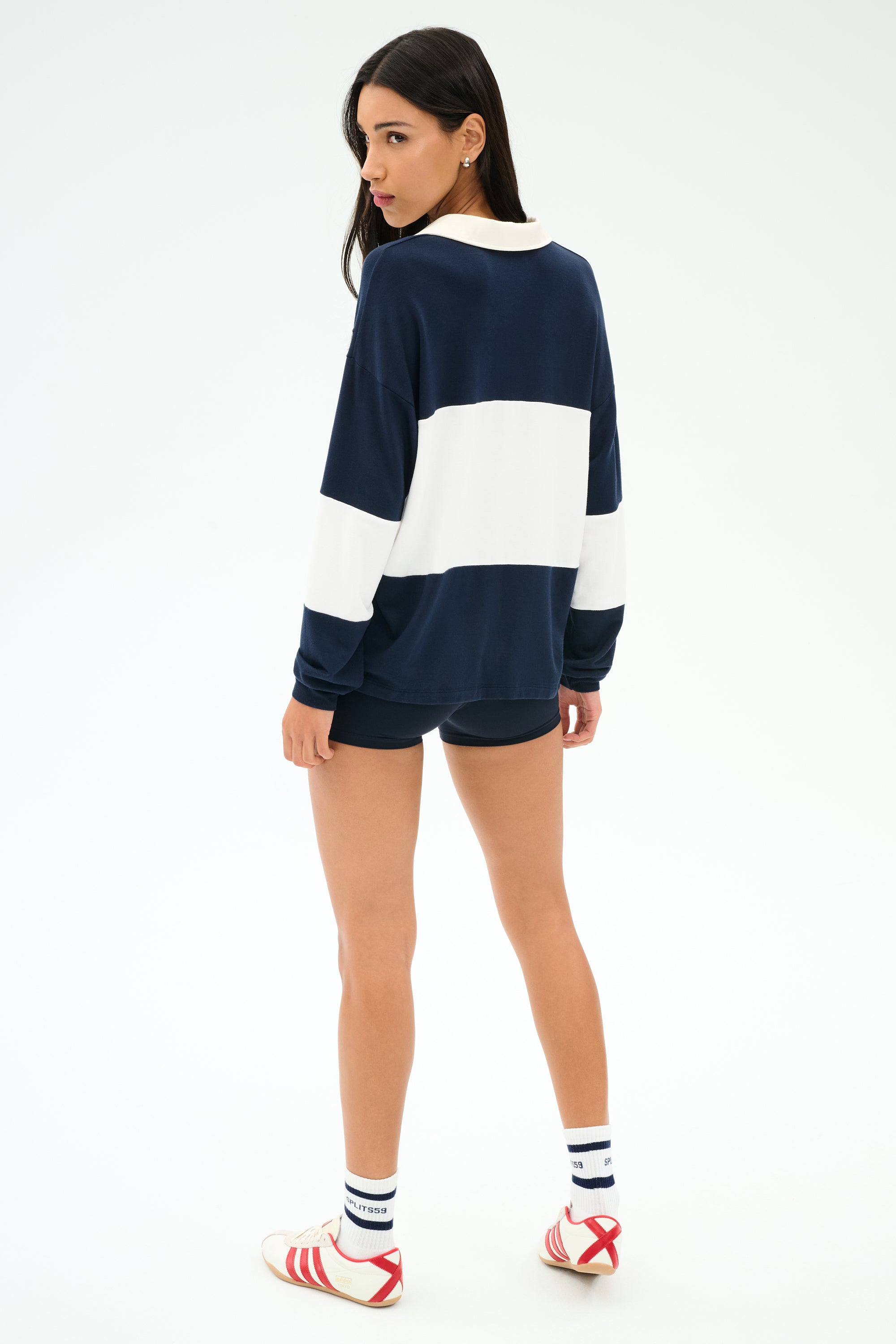 A person stands slightly turned, wearing the SPLITS59 Katie Fleece Rugby in Indigo/White, navy shorts, white socks, and red-striped athletic shoes against a plain light background.