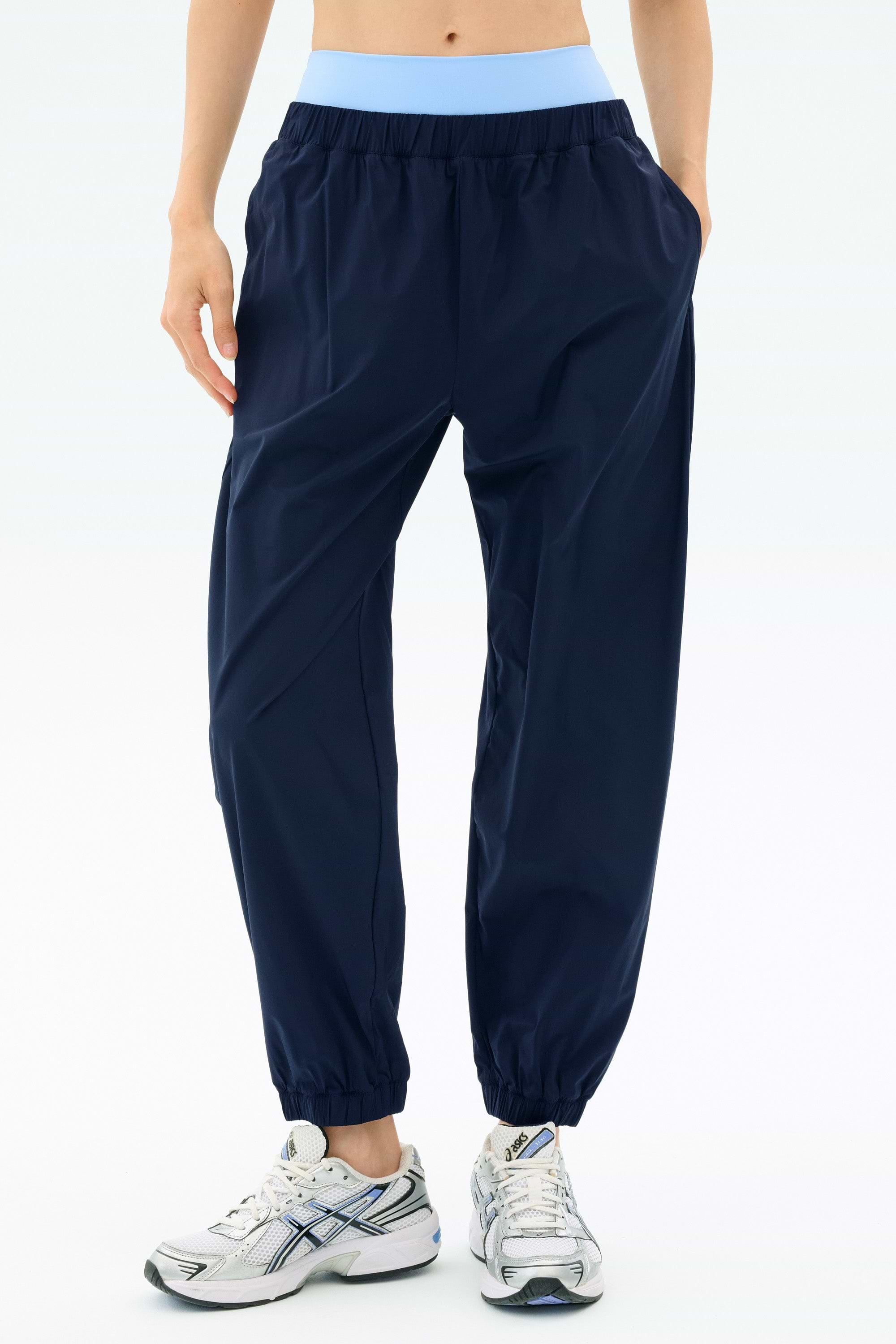 Model wearing SPLITS59 Megan Woven Nylon Jogger in Indigo, featuring elastic cuffs and a light blue waistband, paired with white sneakers and hands in pockets, standing against a plain background.