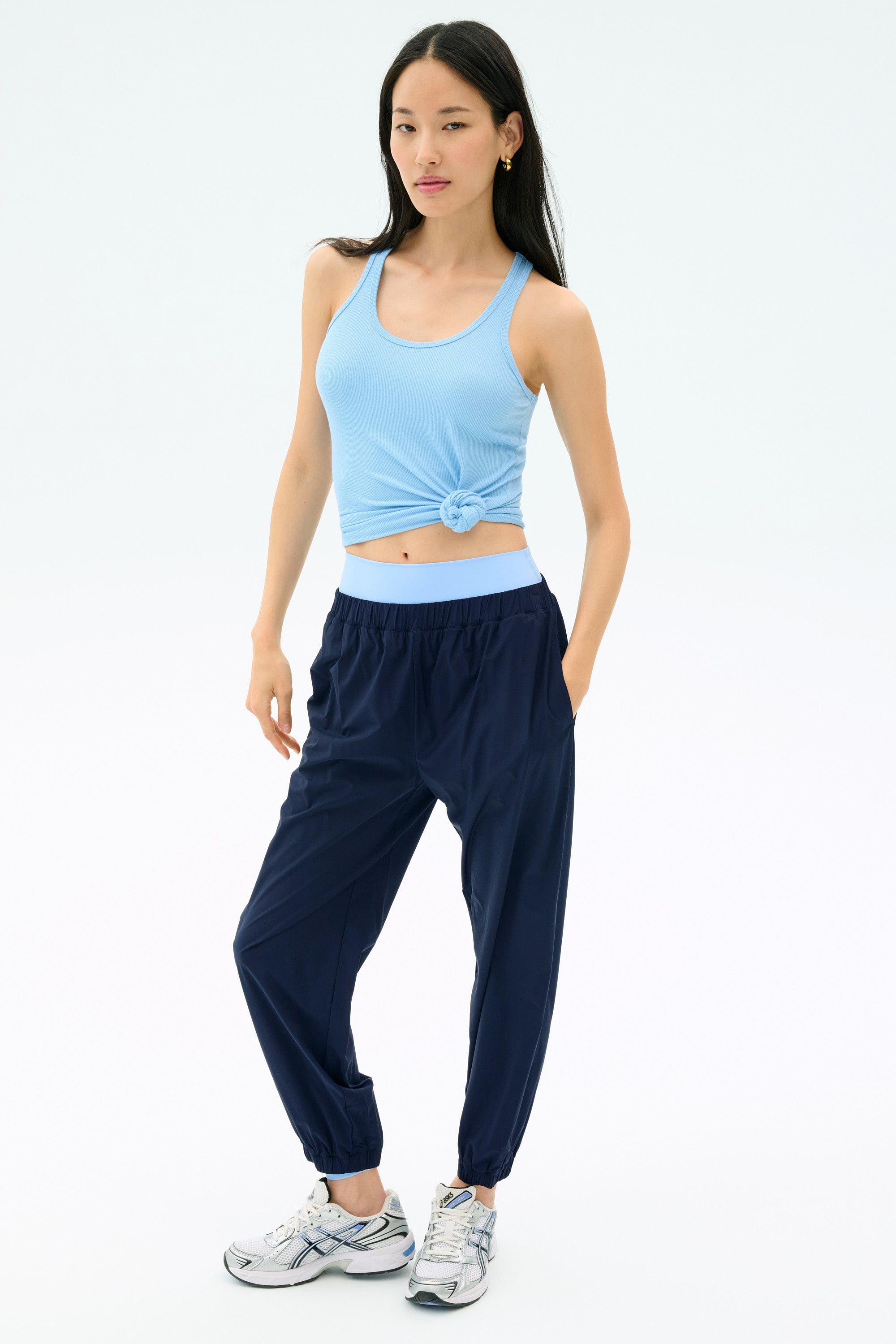 A woman faces forward against a white background, wearing the SPLITS59 Megan Woven Nylon Jogger in Indigo, a light blue knotted tank top, and gray sneakers.