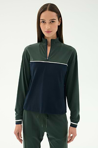 A woman with straight brown hair wears the SPLITS59 Millie Airweight Half Zip in green and navy with matching green pants, standing against a plain light background.