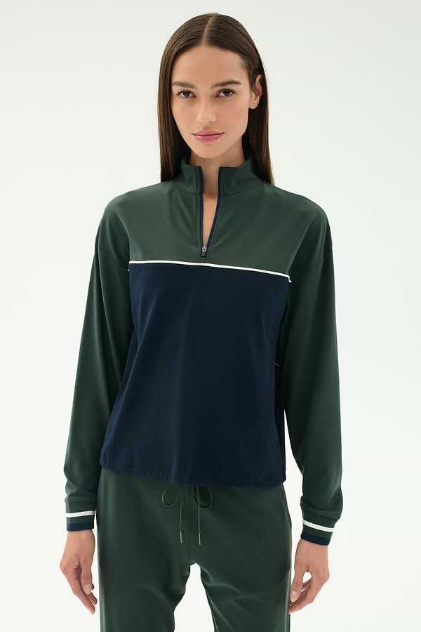 A woman with straight brown hair wears the SPLITS59 Millie Airweight Half Zip in green and navy with matching green pants, standing against a plain light background.