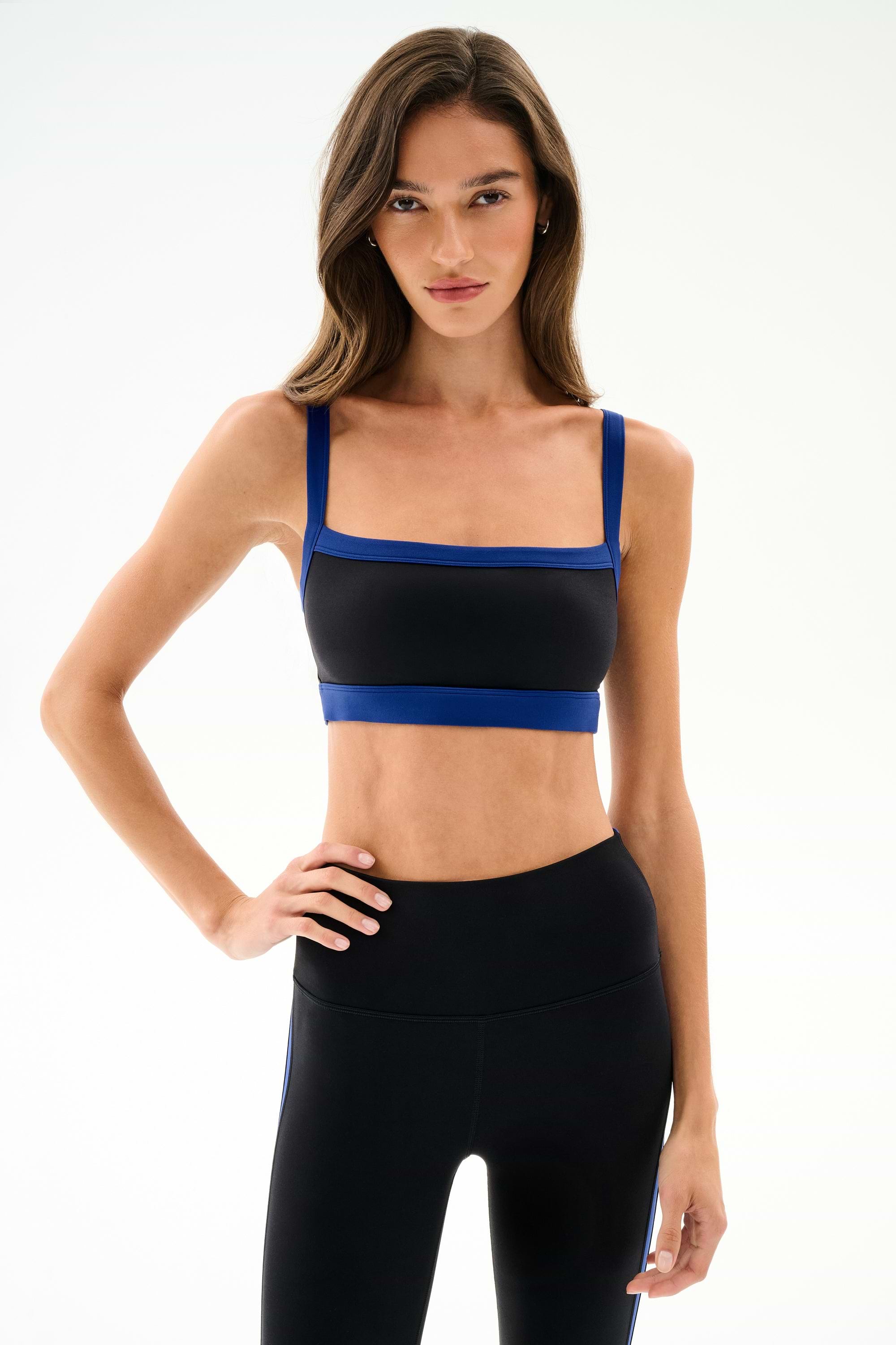 Woman with long brown hair wears the Monah Rigor Bra - Black/Cobalt by SPLITS59, standing with one hand on her hip against a plain light background.