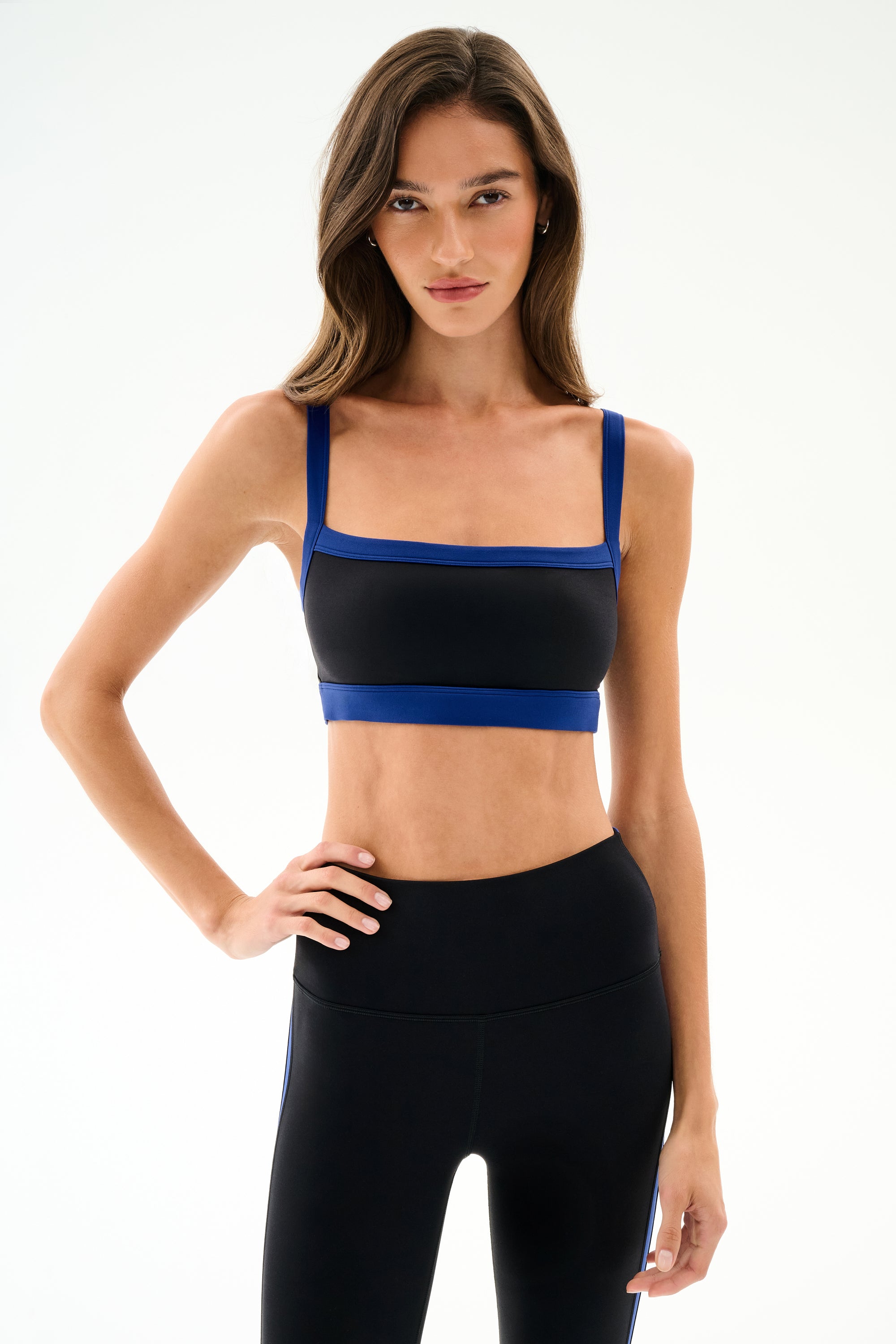 Woman with long brown hair wears the Monah Rigor Bra - Black/Cobalt by SPLITS59, standing with one hand on her hip against a plain light background.