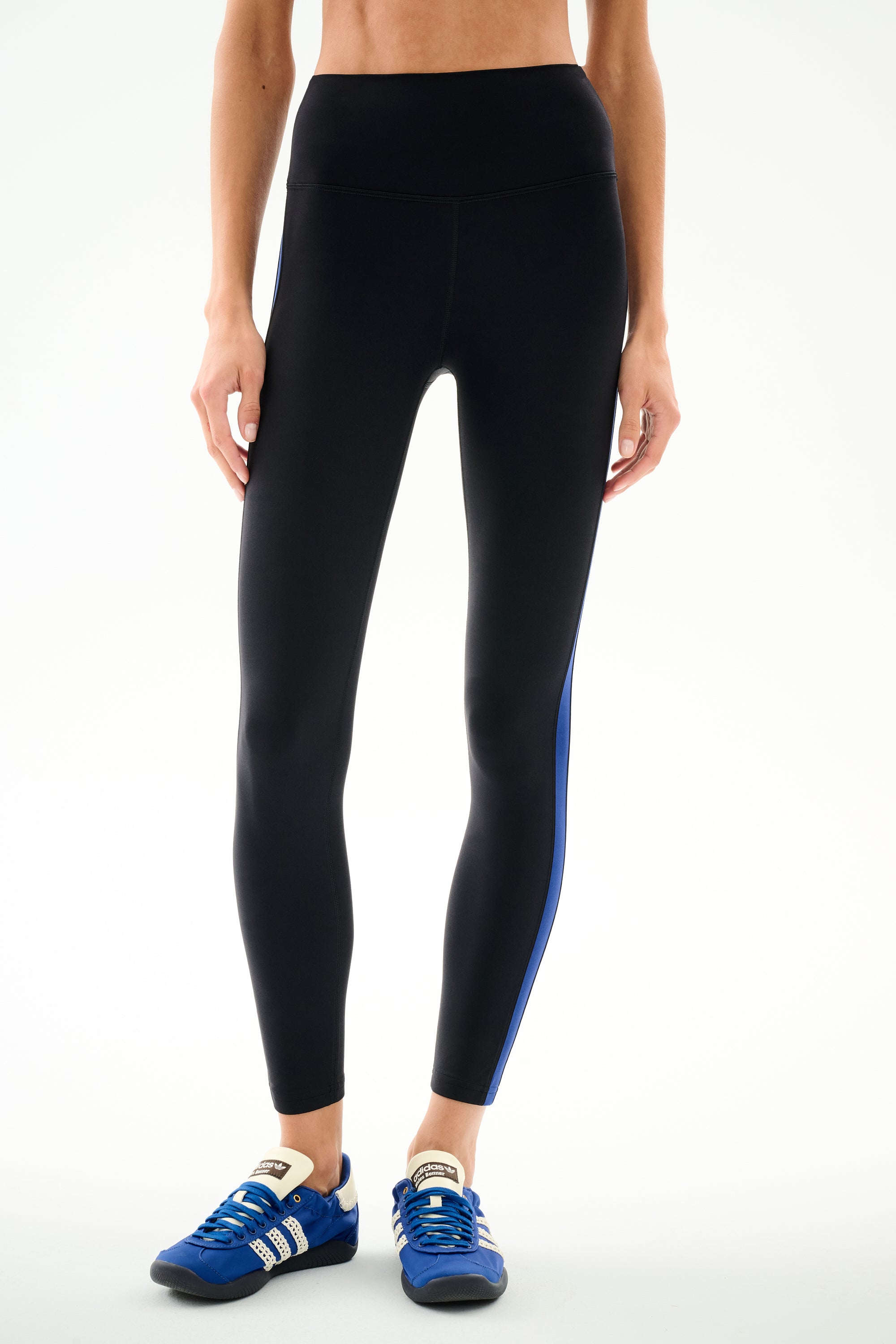 Wearing SPLITS59 Clare High Waist Rigor 7/8 - Black/Cobalt leggings with a blue side stripe and blue sneakers, standing against a light background.
