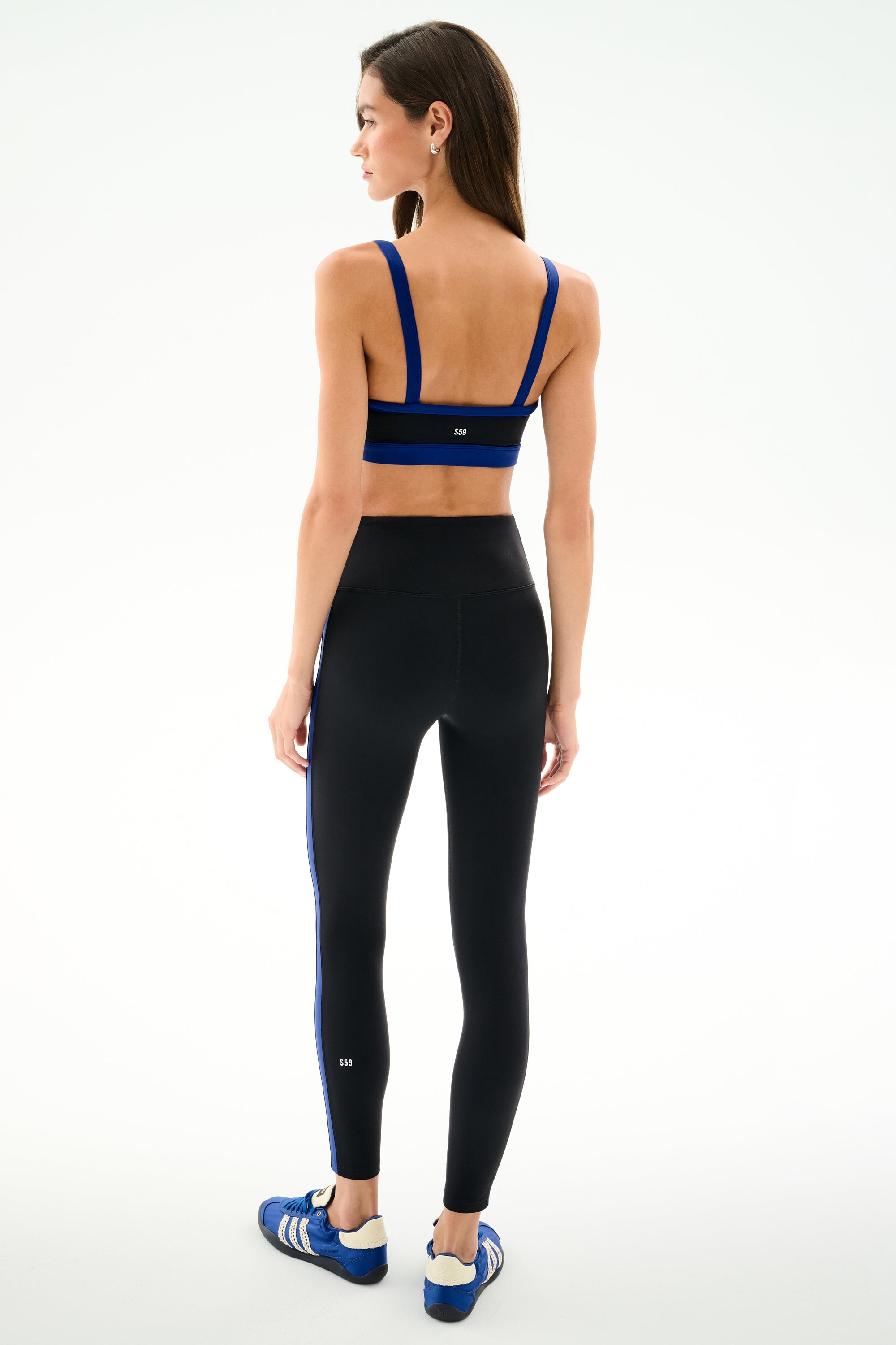 Woman stands with back to camera wearing SPLITS59 Clare High Waist Rigor 7/8 - Black/Cobalt leggings, blue-striped sports bra, and blue athletic shoes on a plain background.