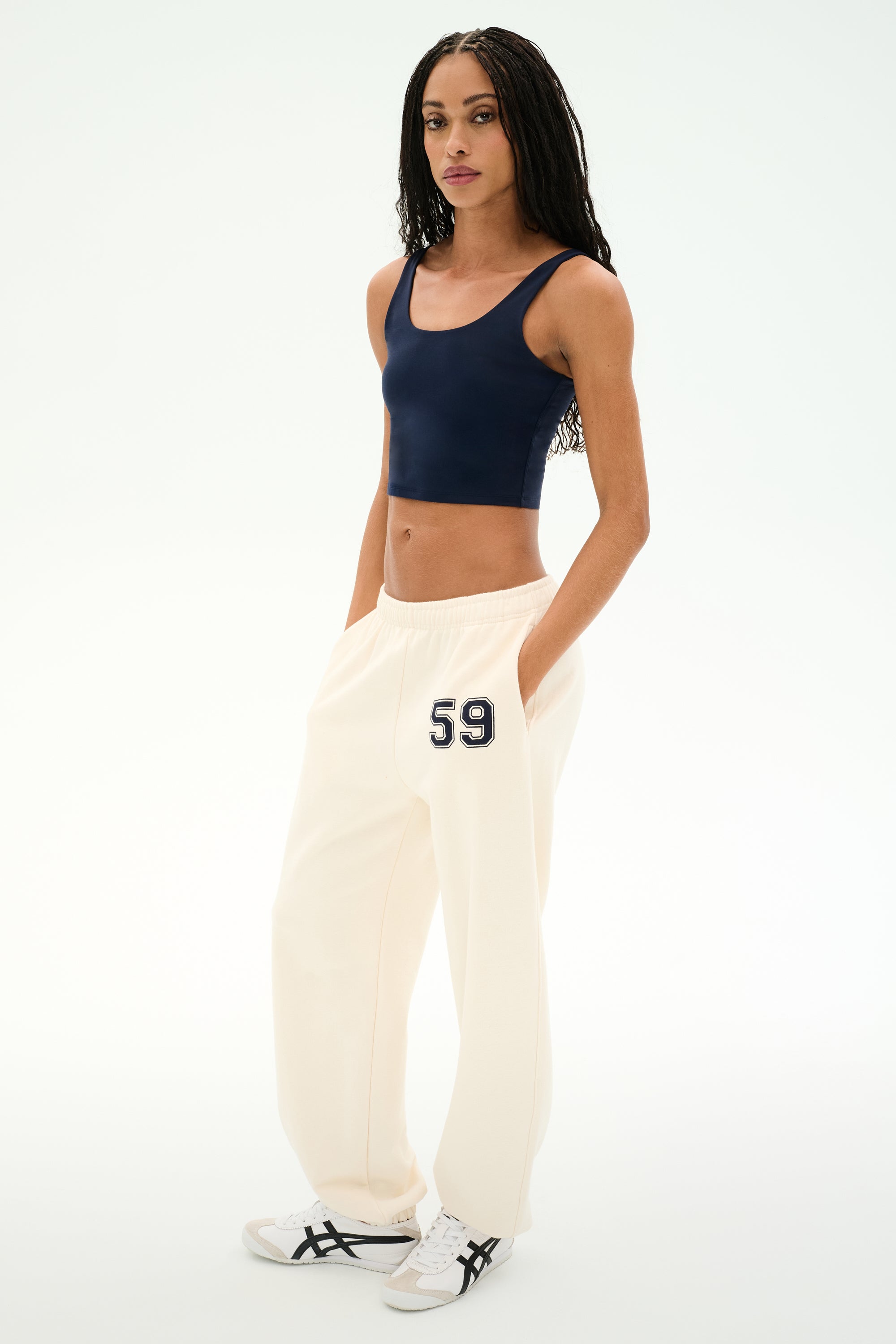 A woman stands against a white background wearing a navy crop top, white sneakers with black stripes, and SPLITS59 Serena French Terry Oversized Sweatpant in Creme/59—crafted from soft cotton and featuring "59" on the hip.