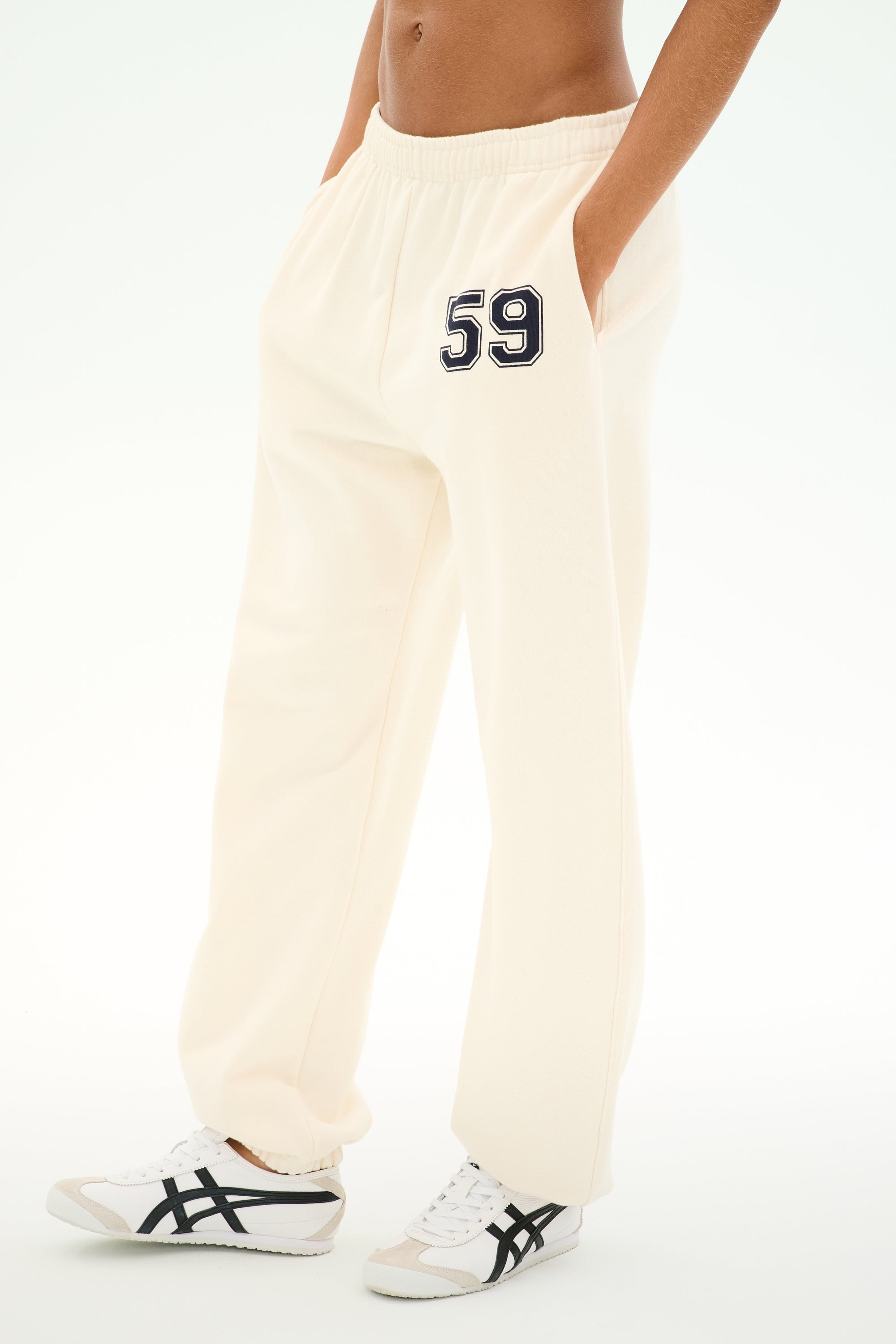 A person wearing the SPLITS59 Serena French Terry Oversized Sweatpant in Creme/59—a pair of 100% cotton cream-colored sweatpants with "59" on the left thigh and pockets—paired with white sneakers featuring black stripes, stands against a plain background.