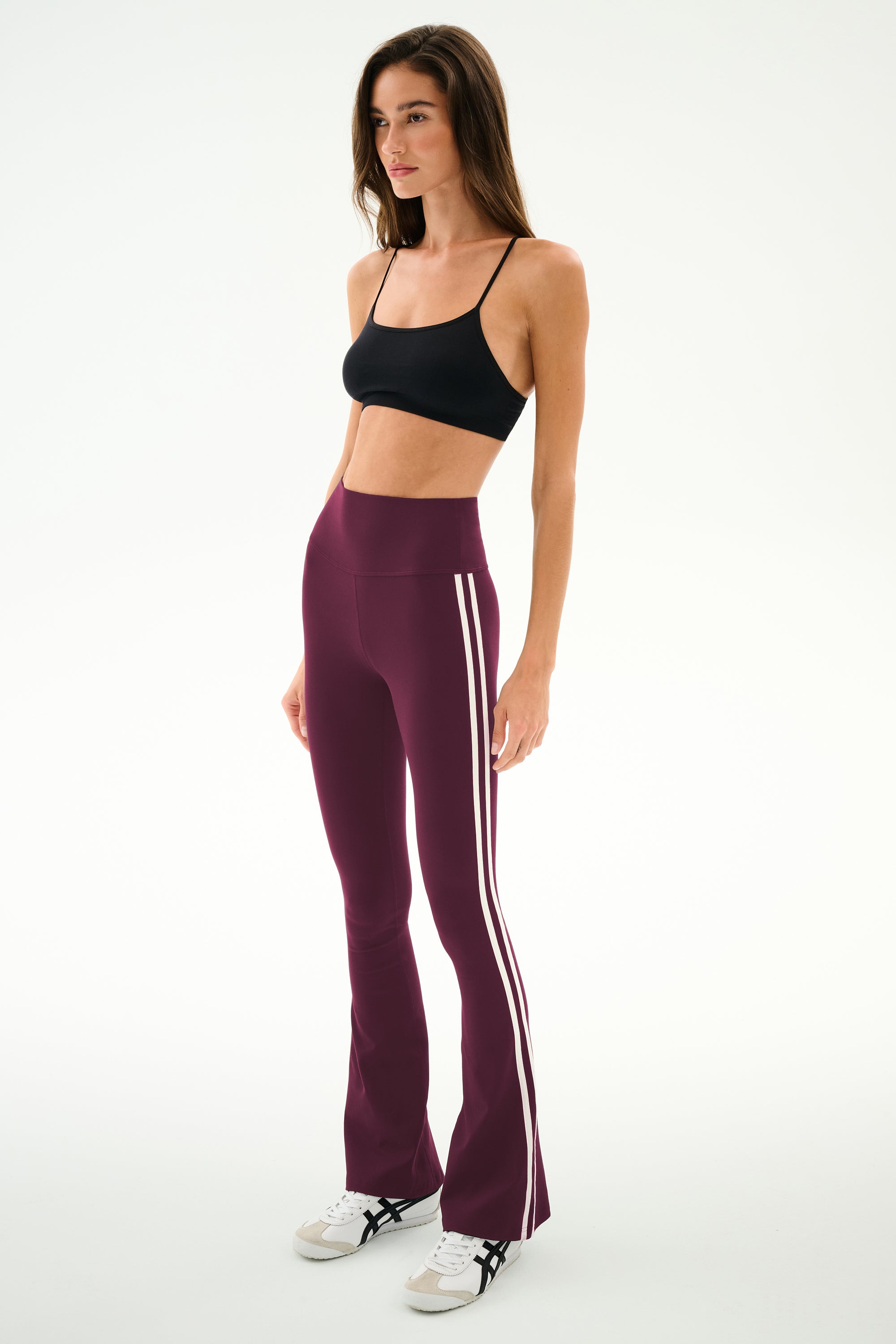 A woman stands against a plain background, wearing the SPLITS59 Raquel High Waist Supplex Flare leggings in Dark Cherry/Creme—a maroon, high-waisted flared design featuring white stripes and crafted from a comfortable nylon-spandex blend. She pairs them with a black sports bra and finishes the look with white sneakers accented by black details.
