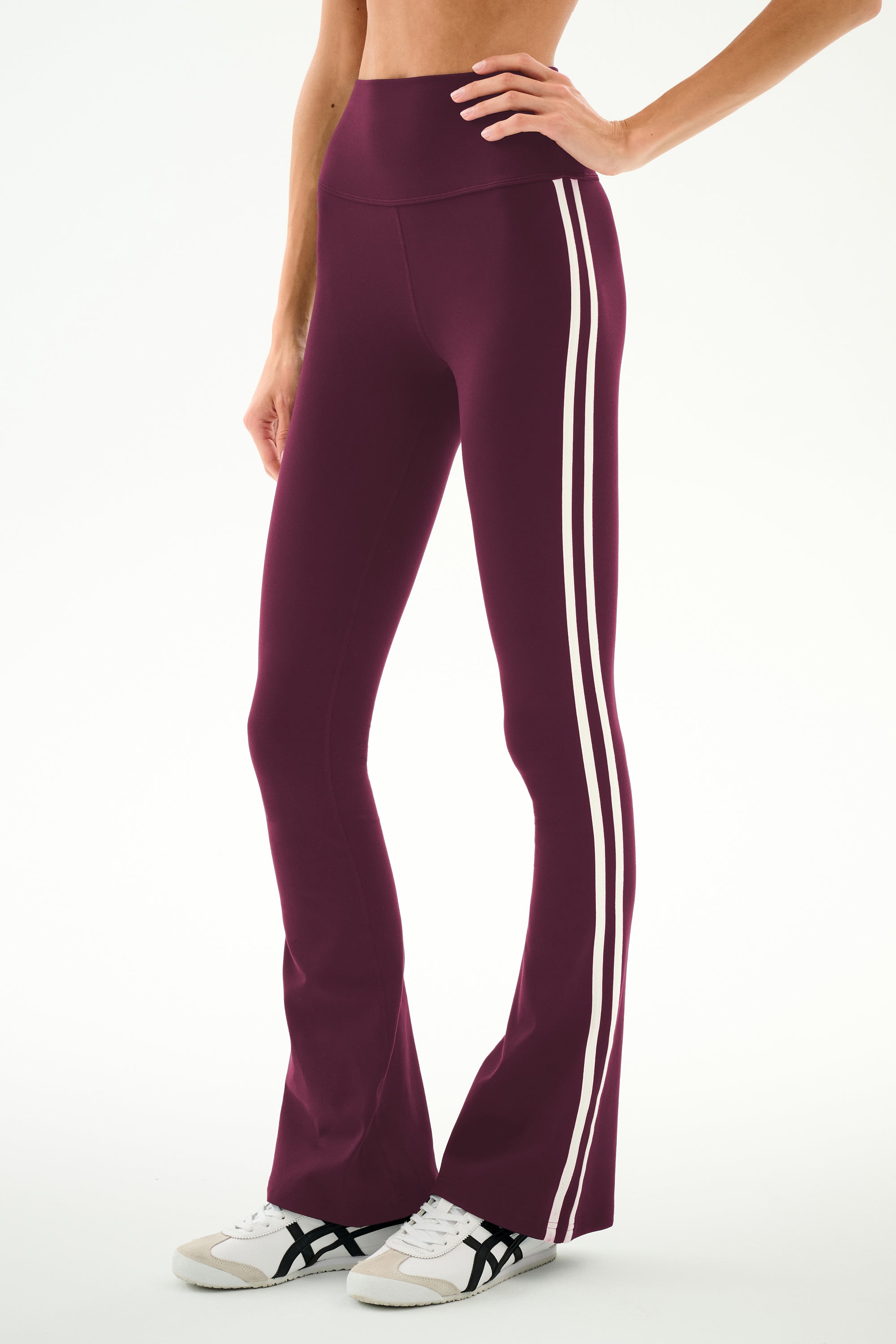 A person wearing the SPLITS59 Raquel High Waist Supplex Flare leggings in Dark Cherry/Creme—a comfortable nylon-spandex blend designed with two white side stripes—paired with white sneakers and standing against a plain background.