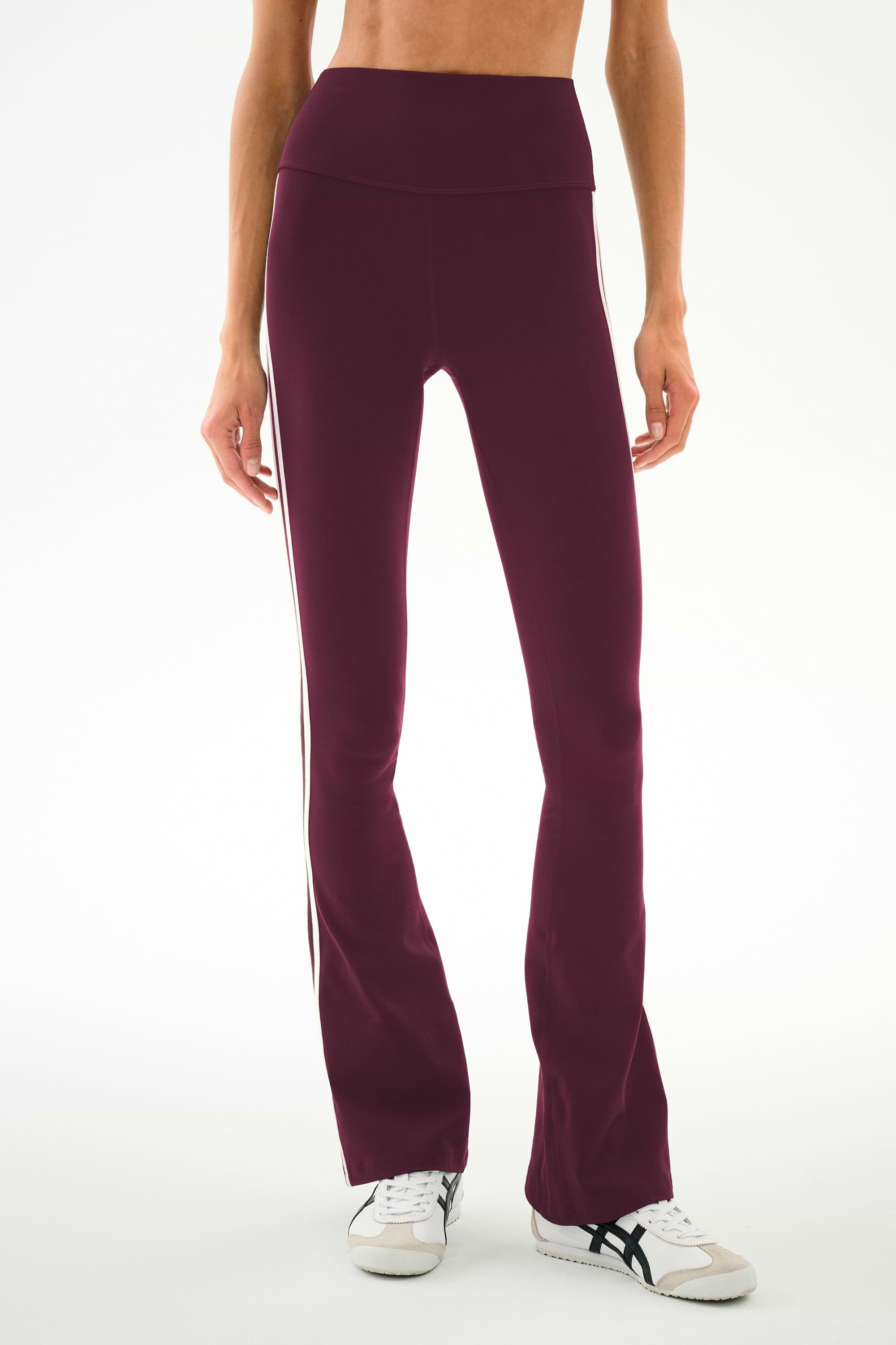 A person wears the SPLITS59 Raquel High Waist Supplex Flare leggings in Dark Cherry/Creme, featuring a high-waisted fit, soft spandex fabric, and white stripe detailing, paired with white sneakers and standing against a plain background.