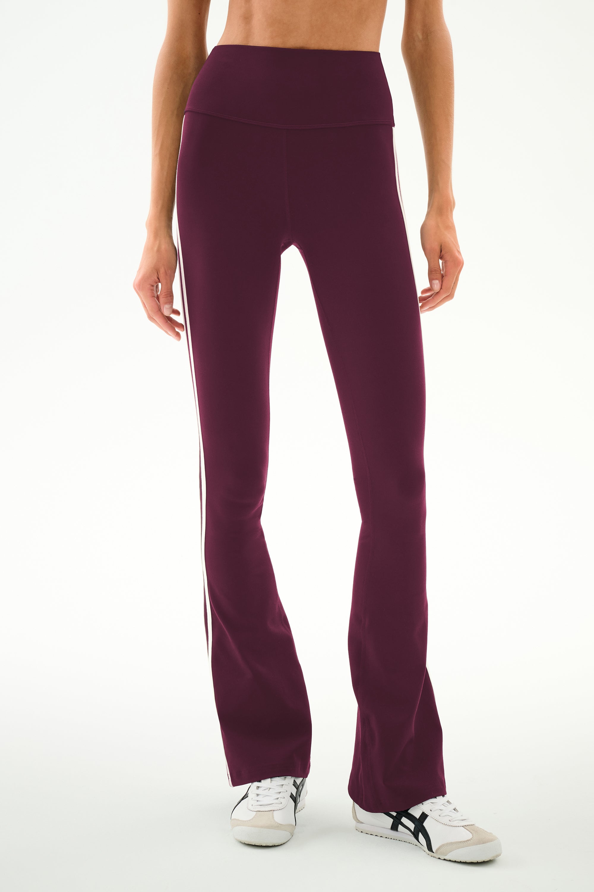 A person wears the SPLITS59 Raquel High Waist Supplex Flare leggings in Dark Cherry/Creme, featuring a high-waisted fit, soft spandex fabric, and white stripe detailing, paired with white sneakers and standing against a plain background.