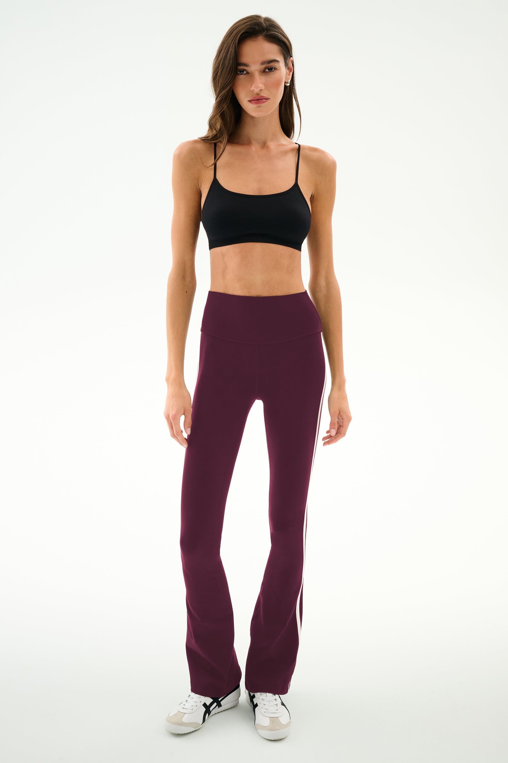 A woman wearing a black nylon-spandex crop top, SPLITS59 Raquel High Waist Supplex Flare pants in Dark Cherry/Creme, and white sneakers stands against a plain white background.