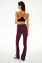 A woman stands facing away, wearing the SPLITS59 Raquel High Waist Supplex Flare leggings in Dark Cherry/Creme—maroon flared leggings with white side stripes—paired with a black sports bra. The leggings are crafted from a comfortable nylon and spandex blend, set against a plain white background.