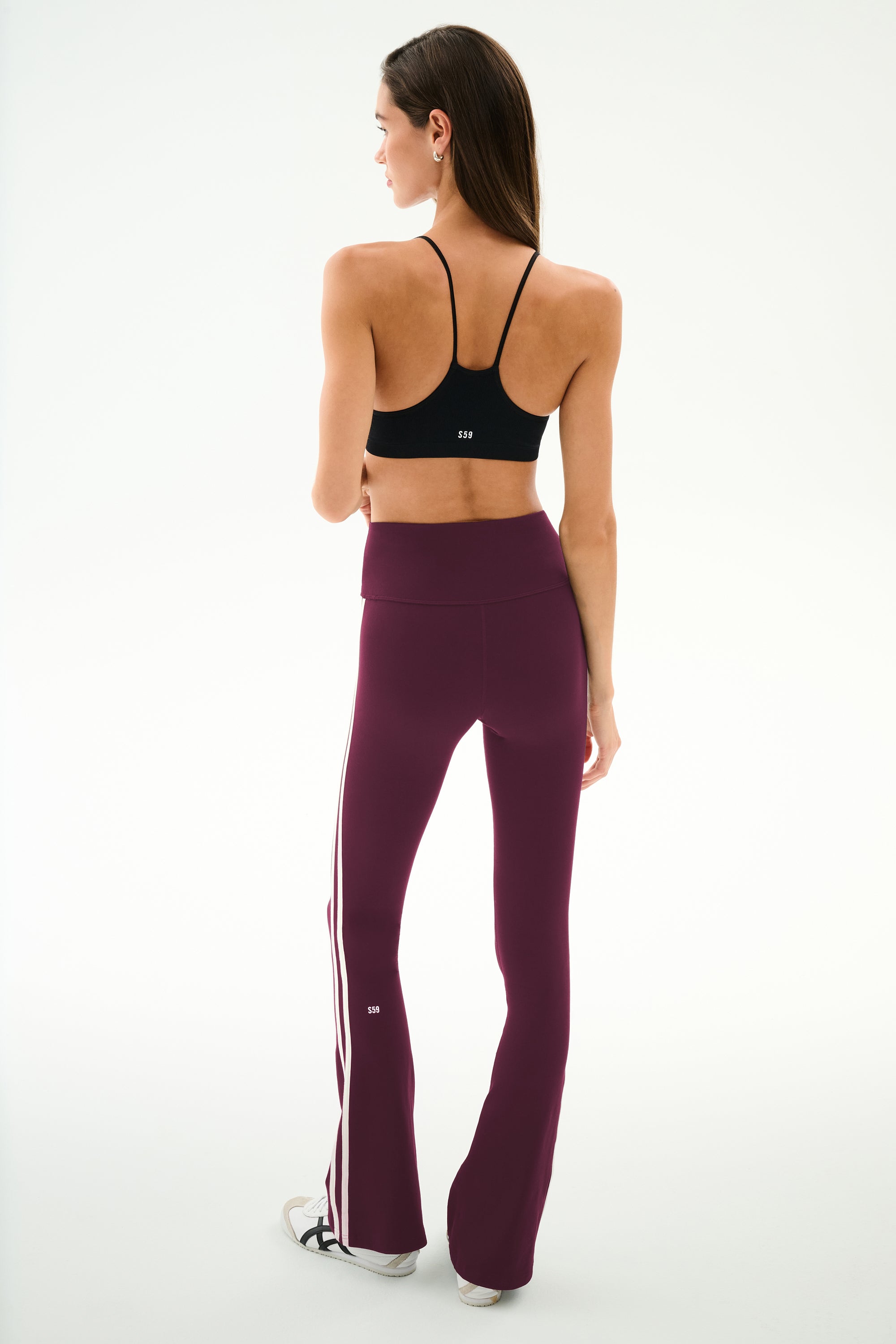 A woman stands facing away, wearing the SPLITS59 Raquel High Waist Supplex Flare leggings in Dark Cherry/Creme—maroon flared leggings with white side stripes—paired with a black sports bra. The leggings are crafted from a comfortable nylon and spandex blend, set against a plain white background.