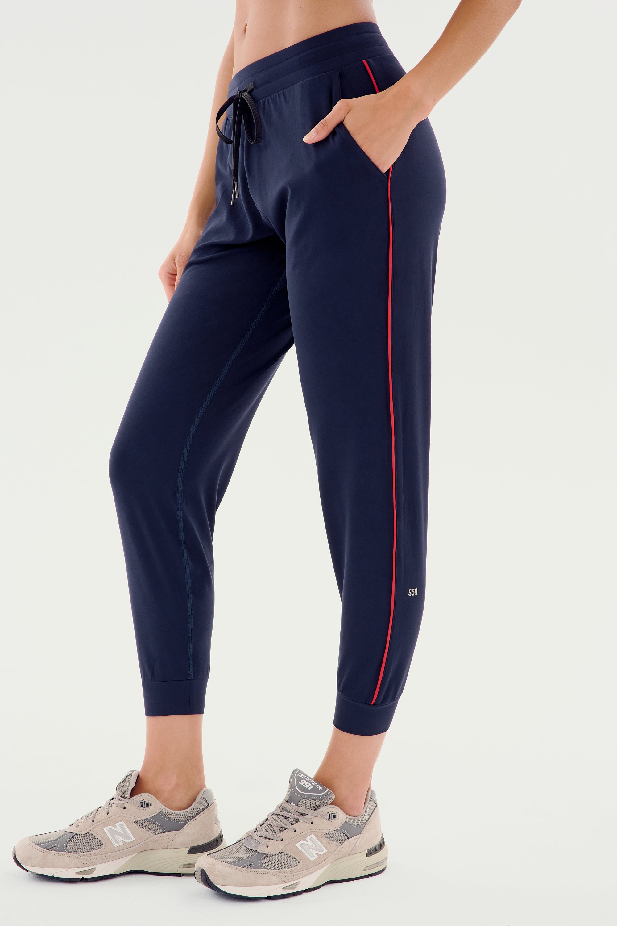 Person wearing SPLITS59 Airweight Jogger 7/8 with Piping - Indigo/Pirate Red and gray sneakers standing on a white background.