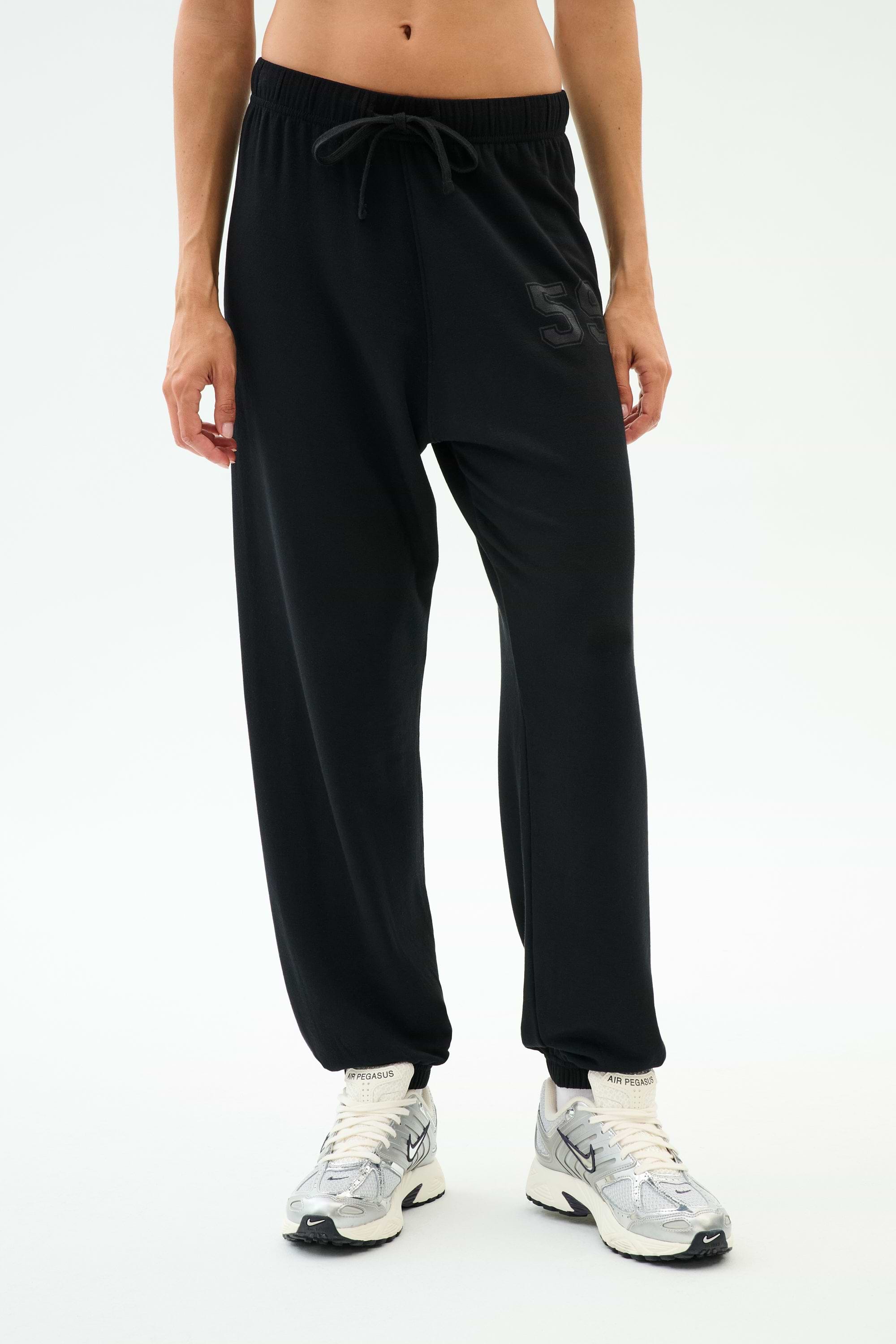A person wears SPLITS59's Andie Oversized Fleece Varsity 59 Sweatpant in black, featuring a "59" on the left thigh, paired with white sneakers—ideal for cool weather workouts—standing against a plain white background.