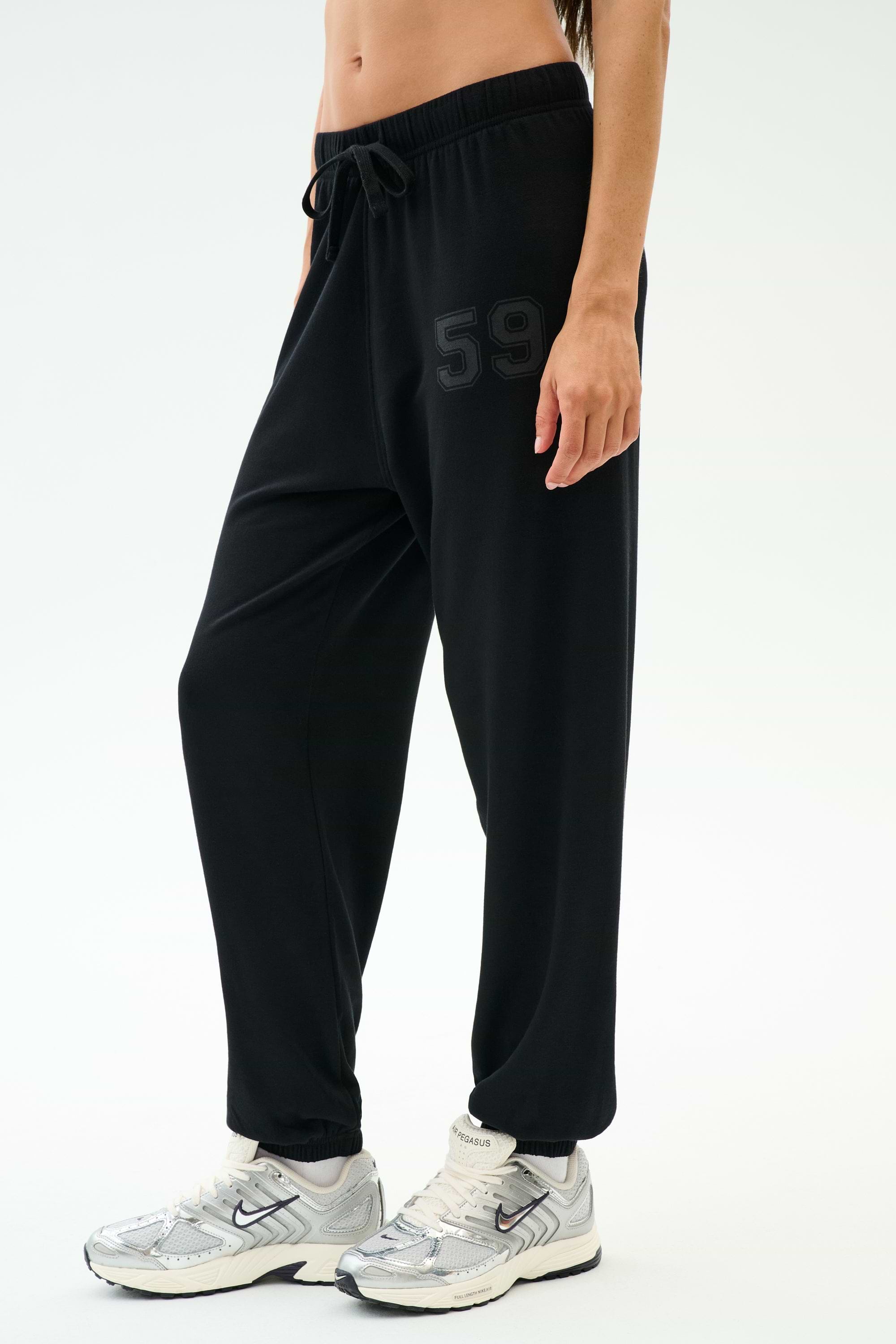 A person wears the SPLITS59 Andie Oversized Fleece Varsity 59 Sweatpant - Black/59, featuring a "59" print on the left thigh, paired with white sneakers and a crop top—perfect for cool weather workouts—against a white background.