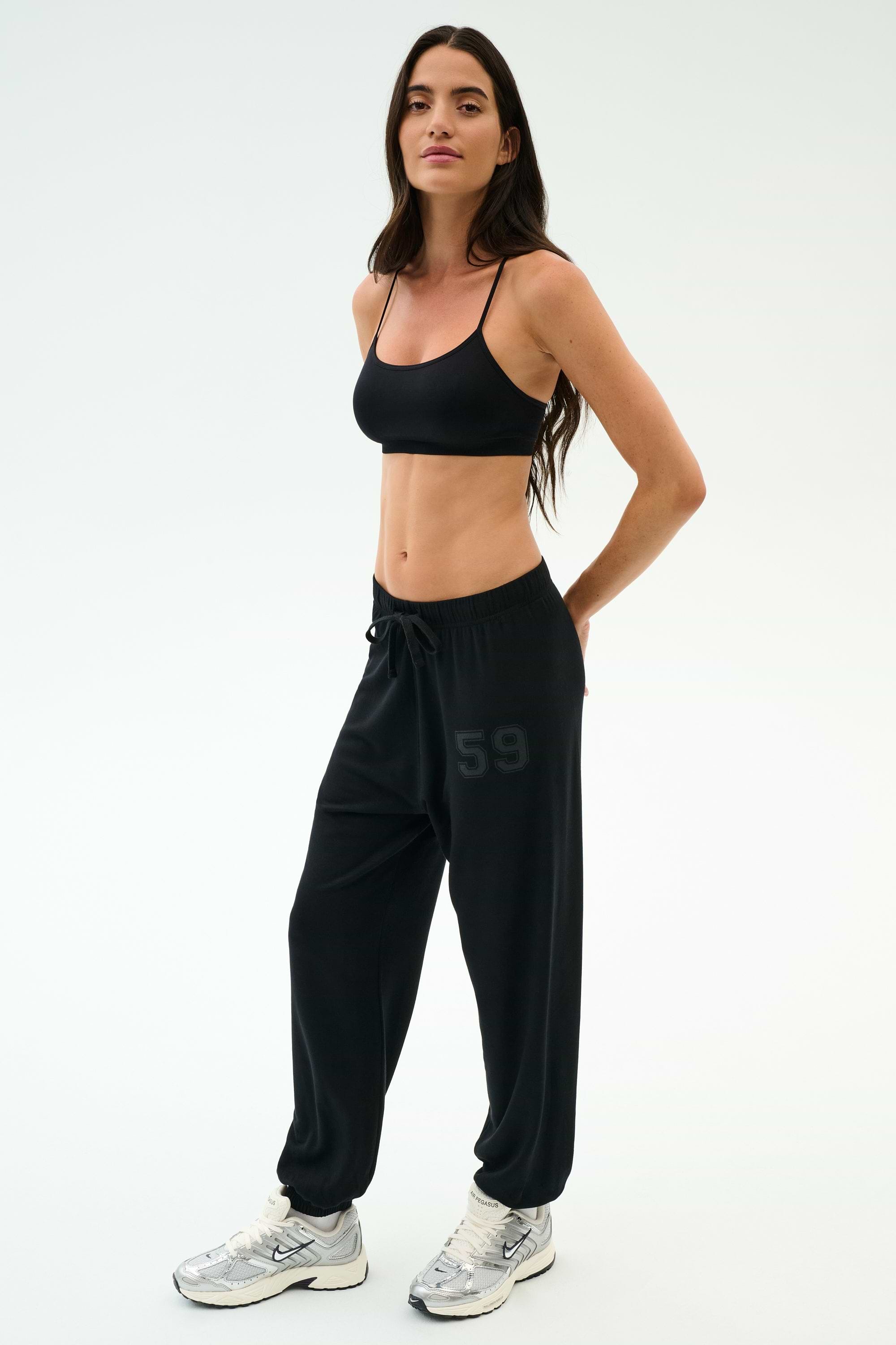 A woman stands in a studio wearing a black sports bra, white sneakers, and SPLITS59’s Andie Oversized Fleece Varsity 59 Sweatpant in Black/59—ultra luxe fleece with "59" on the leg—perfect for cool weather workouts.