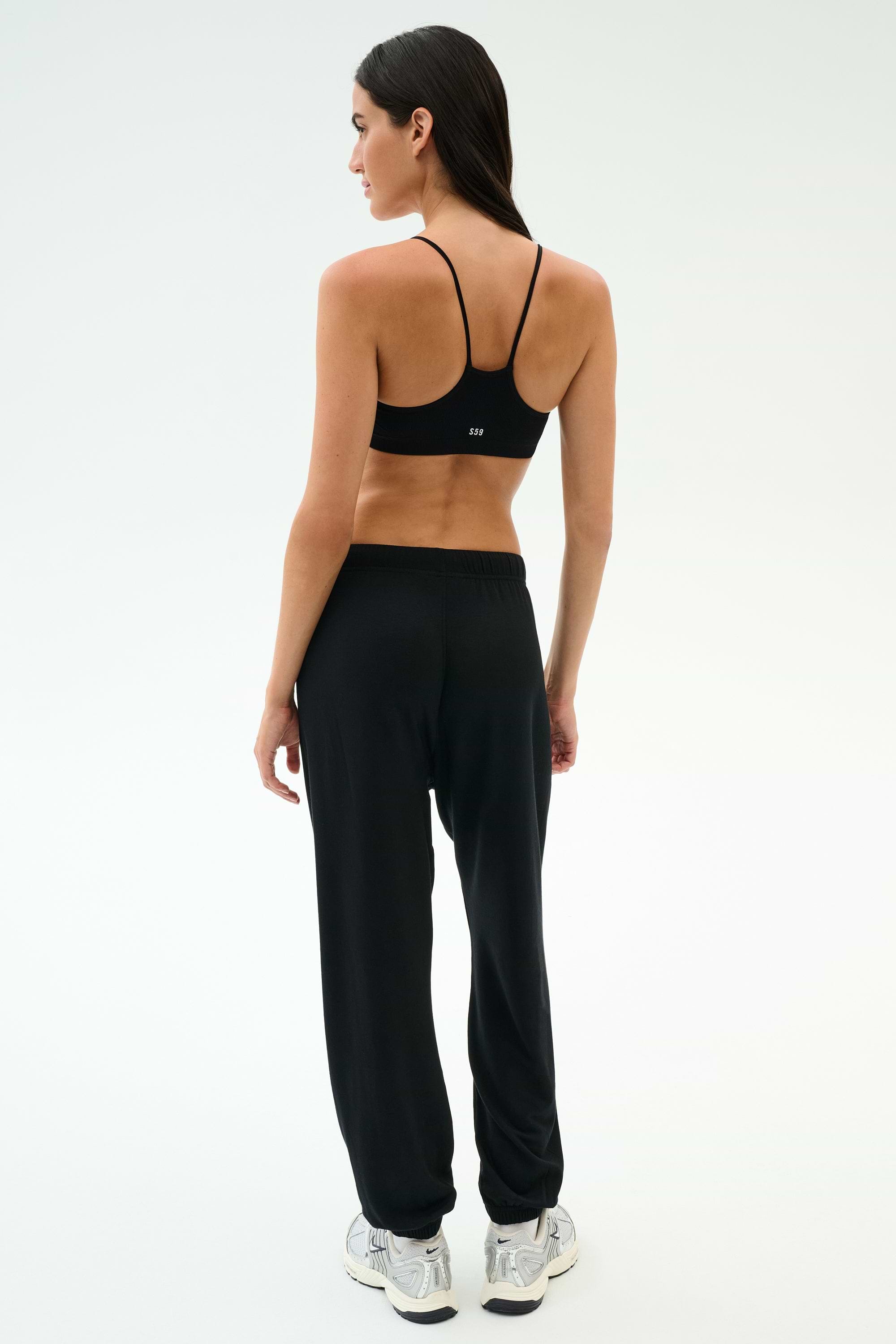 A woman stands with her back to the camera in SPLITS59’s Andie Oversized Fleece Varsity 59 Sweatpant - Black/59, paired with a black sports bra and white sneakers, ideal for cool weather workouts against a plain light background.