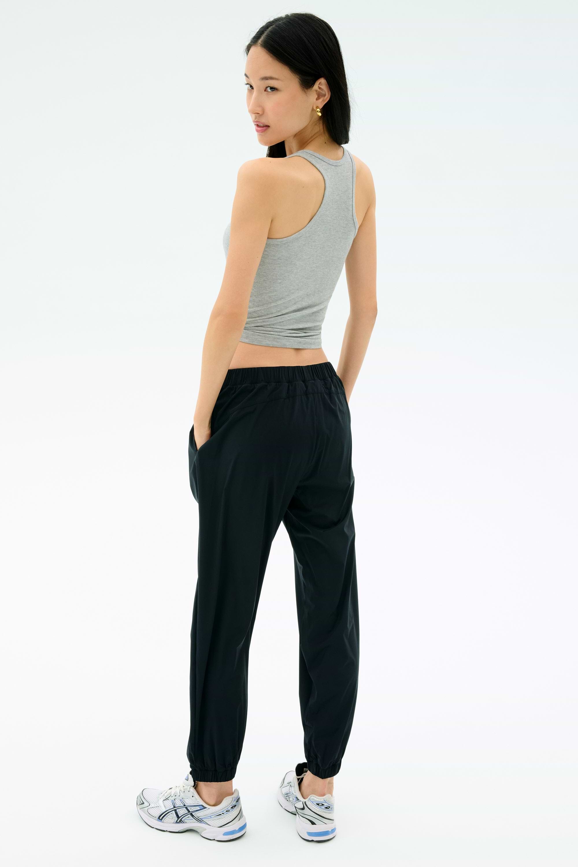 A woman stands with her back turned, wearing the Splits59 Ashby Rib Tank in Lt. Heather Grey, black jogger pants, and white sneakers, hands in pockets against a plain background.