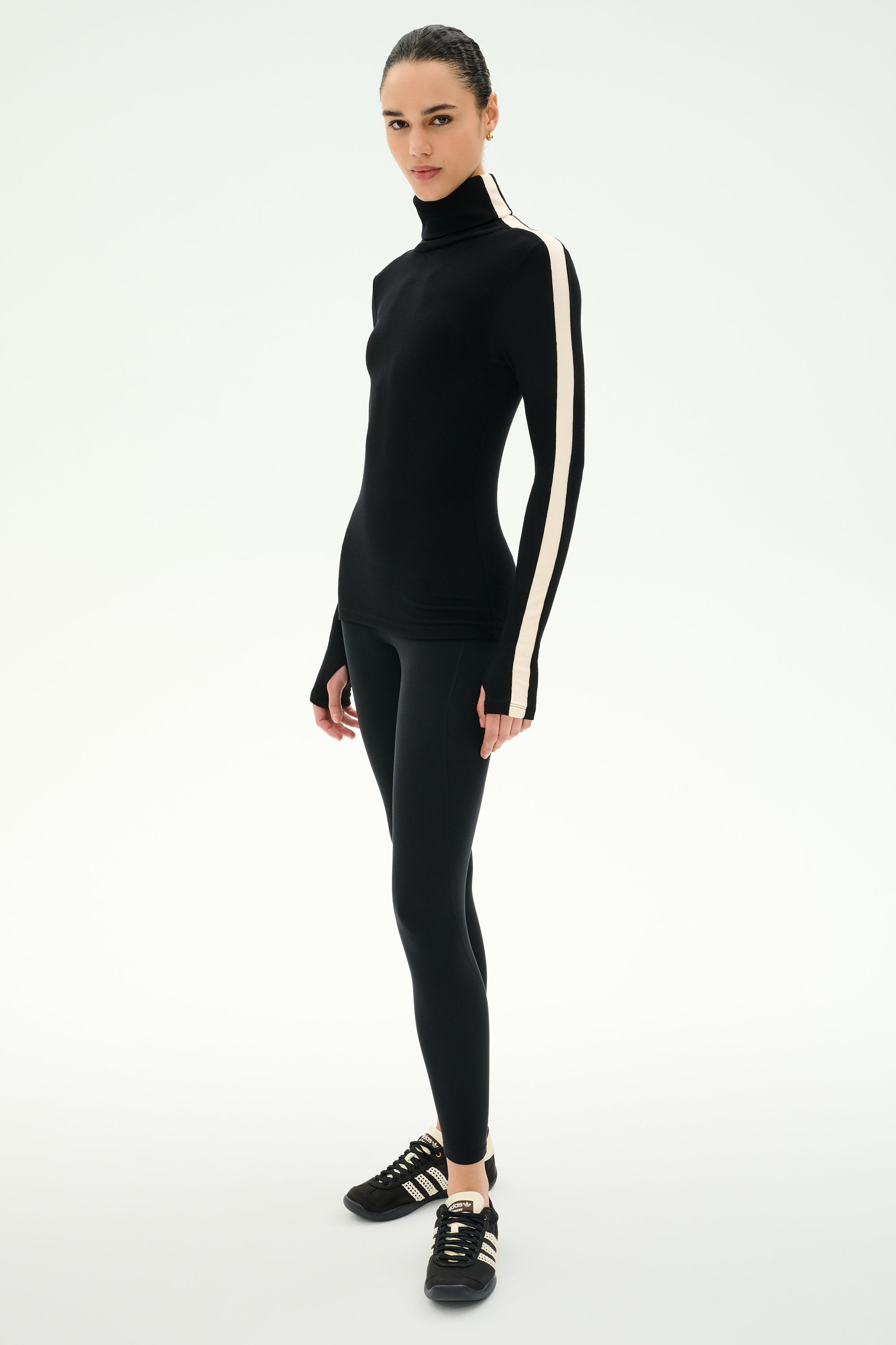 A person wearing the SPLITS59 Jackson Rib Full Length Turtleneck in Black/Creme, which features a white stripe on the sleeve, paired with black leggings and black athletic shoes, standing against a white background.