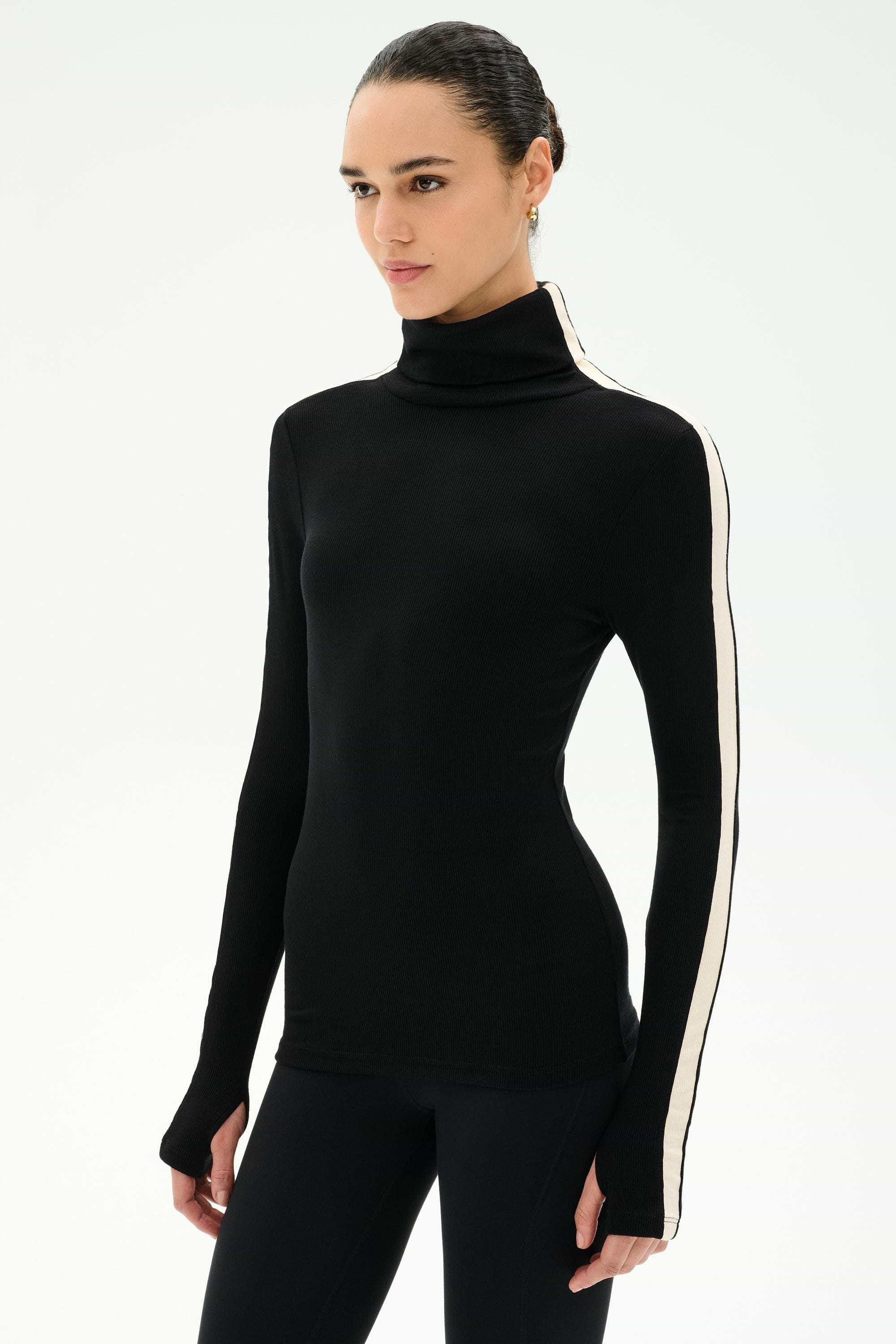 A woman wears the SPLITS59 Jackson Rib Full Length Turtleneck in Black/Creme, featuring a white stripe down the sleeve, as she stands against a plain light background.