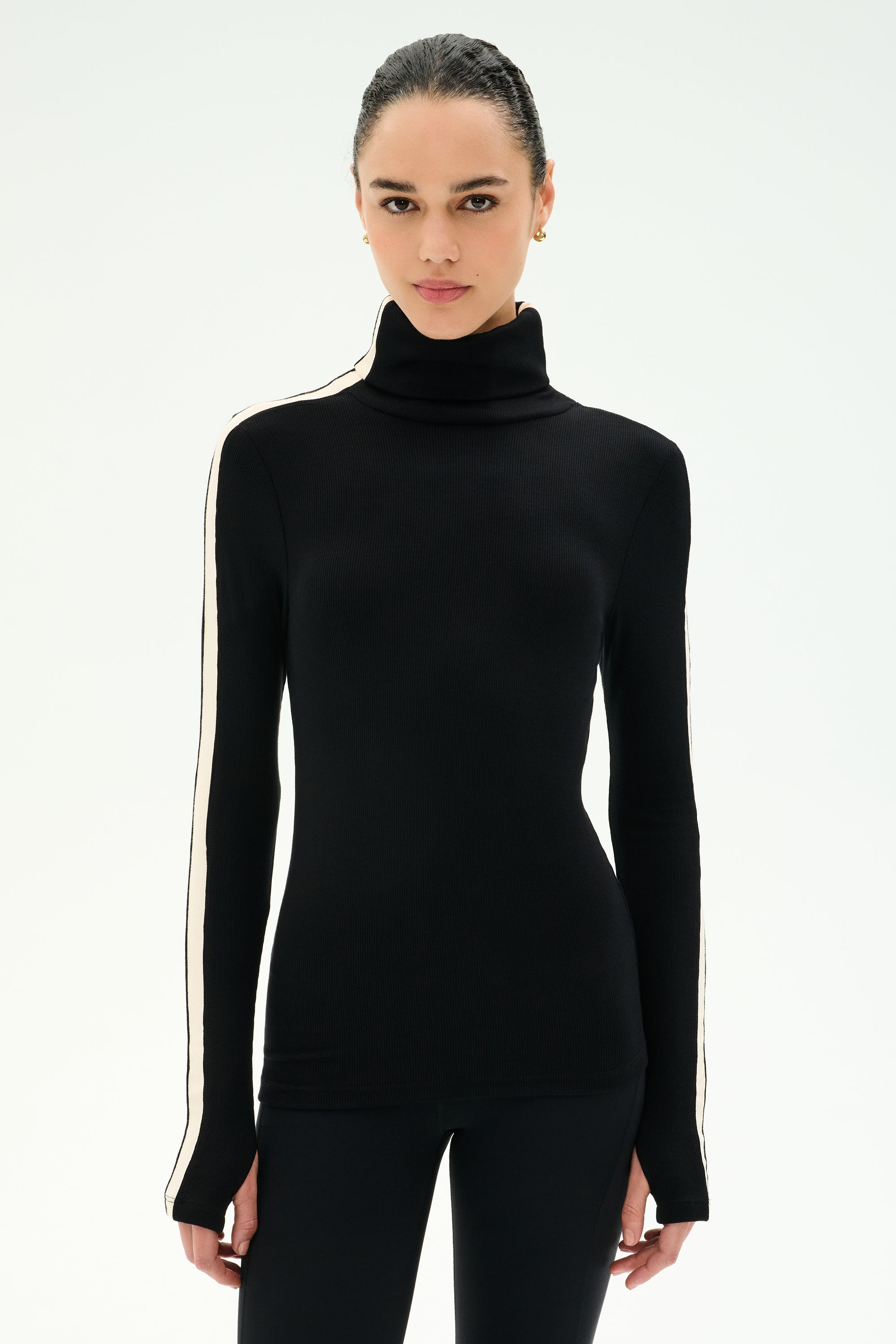 Wearing the SPLITS59 Jackson Rib Full Length Turtleneck in Black/Creme—a black sweater featuring creme stripes along the sleeves—a person stands facing forward against a plain white background.