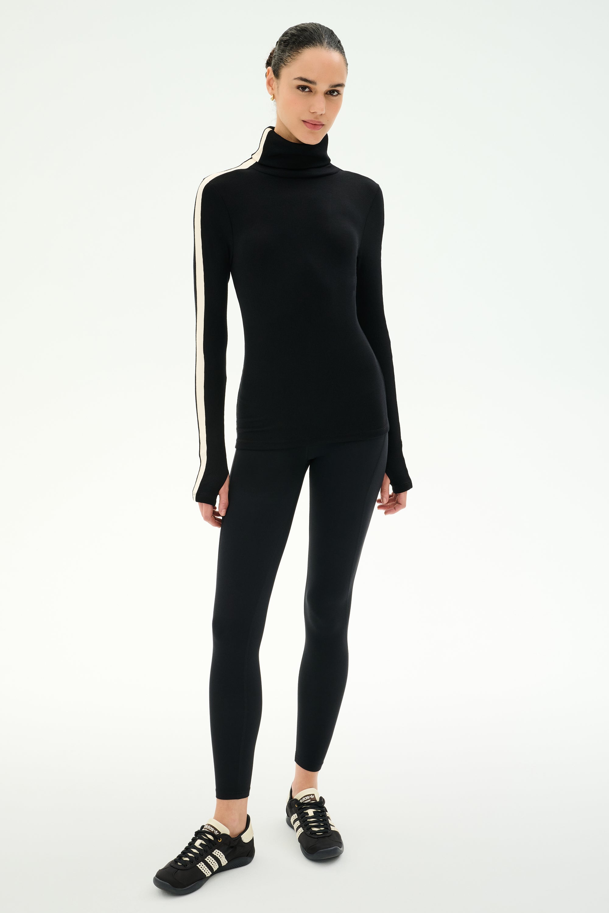 A woman stands against a plain white background wearing the SPLITS59 Jackson Rib Full Length Turtleneck in Black/Creme—a sleek black baby rib turtleneck with creme stripes on the sleeves—paired with black leggings and black sneakers featuring white stripes.