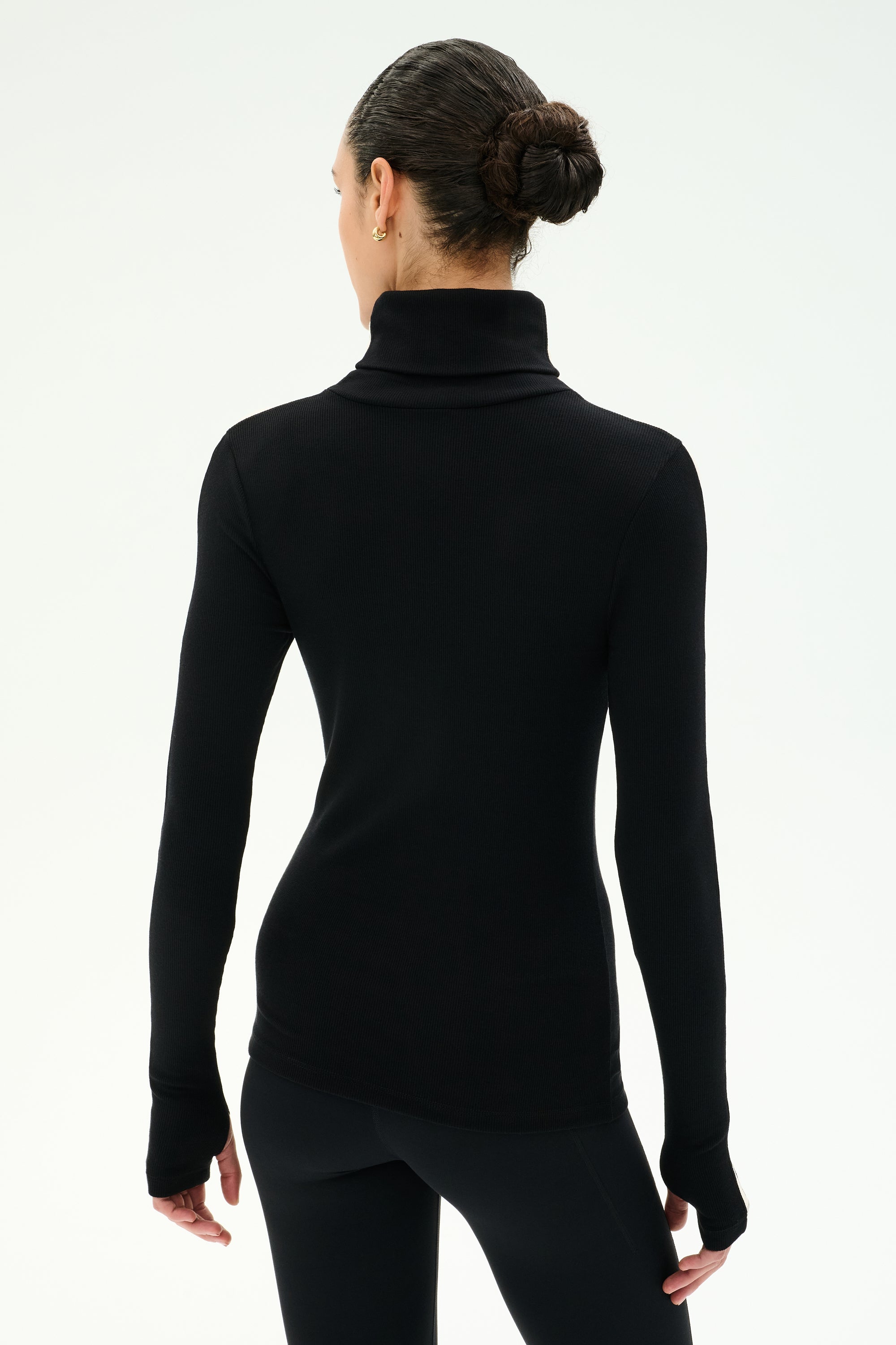 A person with dark hair in a bun is seen from behind, wearing the SPLITS59 Jackson Rib Full Length Turtleneck in Black/Creme and black pants, standing against a plain white background.