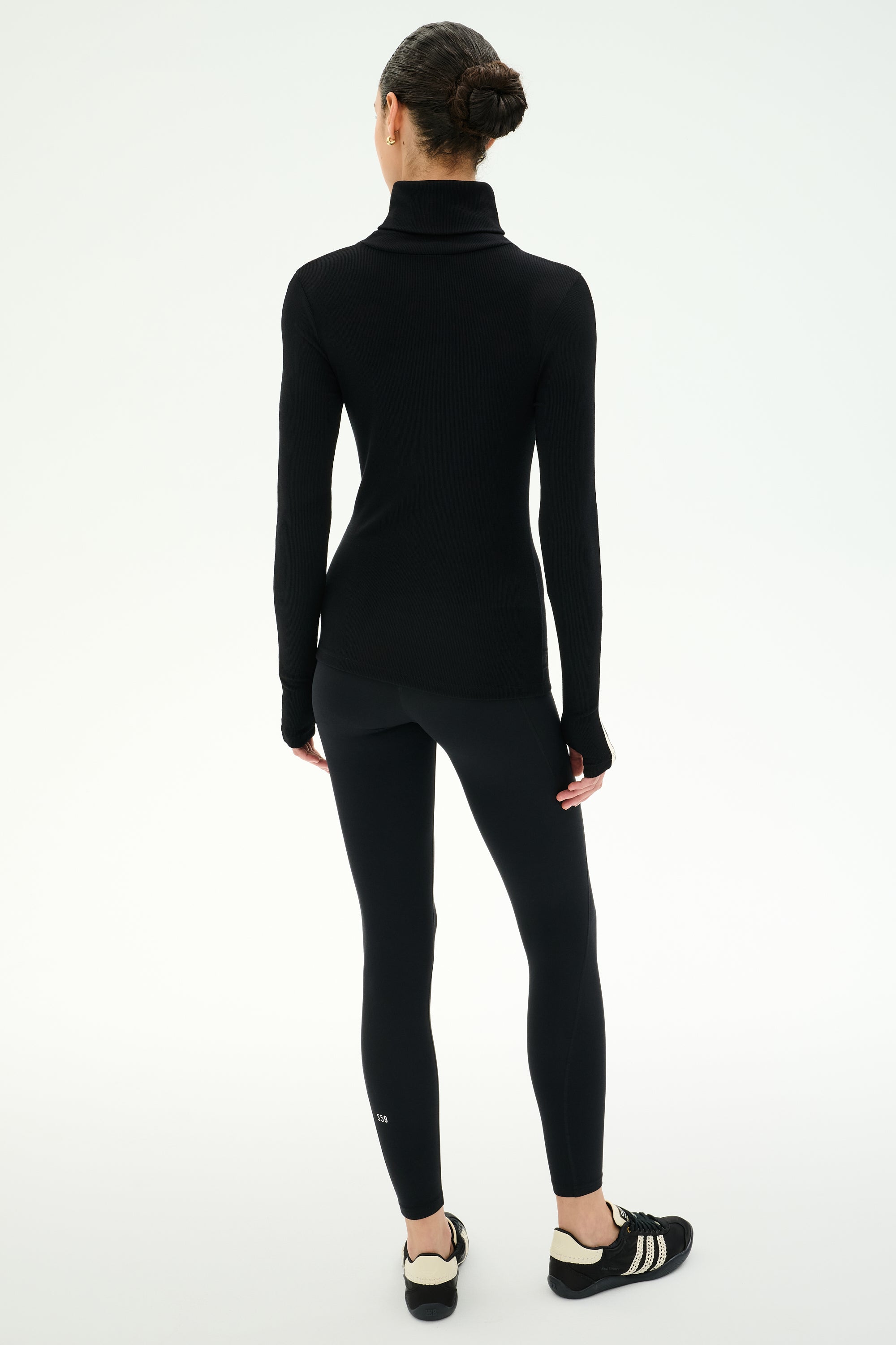 A woman stands facing away, wearing the SPLITS59 Jackson Rib Full Length Turtleneck in Black/Creme, paired with black leggings and black-and-white sneakers against a plain white background.