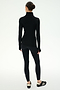 A woman stands facing away, wearing the SPLITS59 Jackson Rib Full Length Turtleneck in Black/Creme, paired with black leggings and black-and-white sneakers against a plain white background.