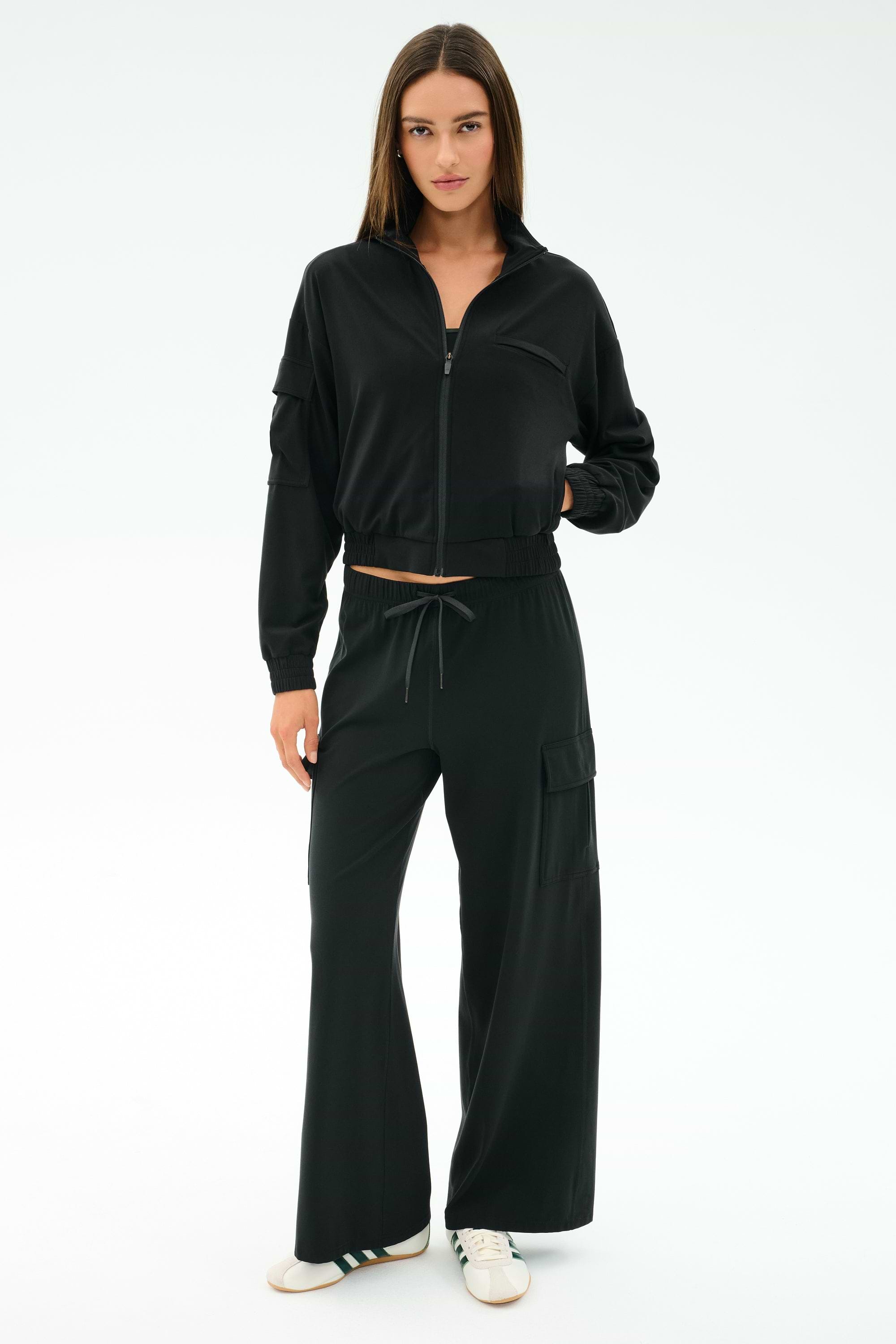 A woman faces forward in a black zip-up jacket and SPLITS59 Alex Airweight Cargo Pant - Black, featuring pockets, paired with white sneakers against a plain white background.