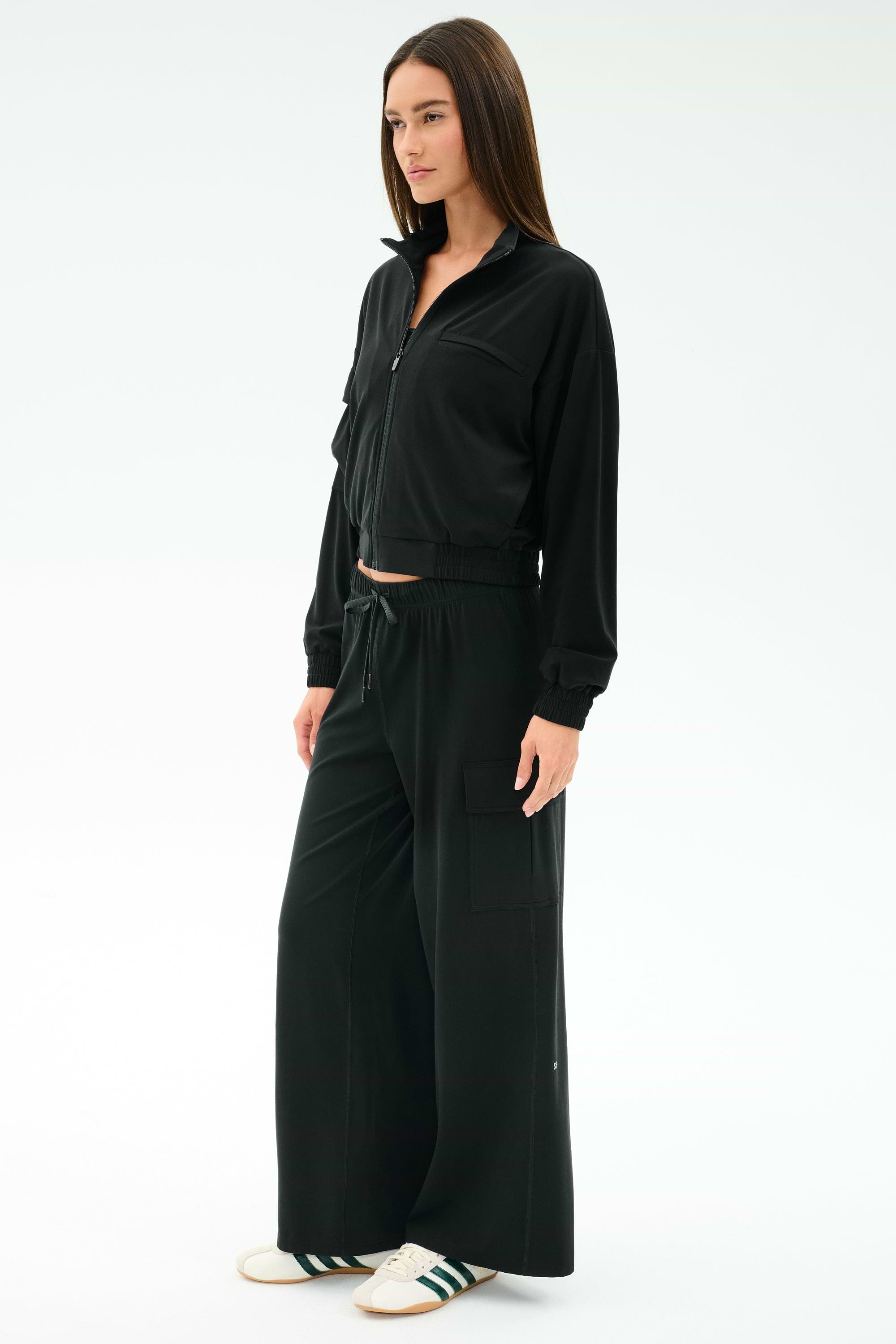 A woman wears the SPLITS59 Peaches Airweight Cargo Jacket in black, paired with matching wide-leg pants and white sneakers with green stripes.