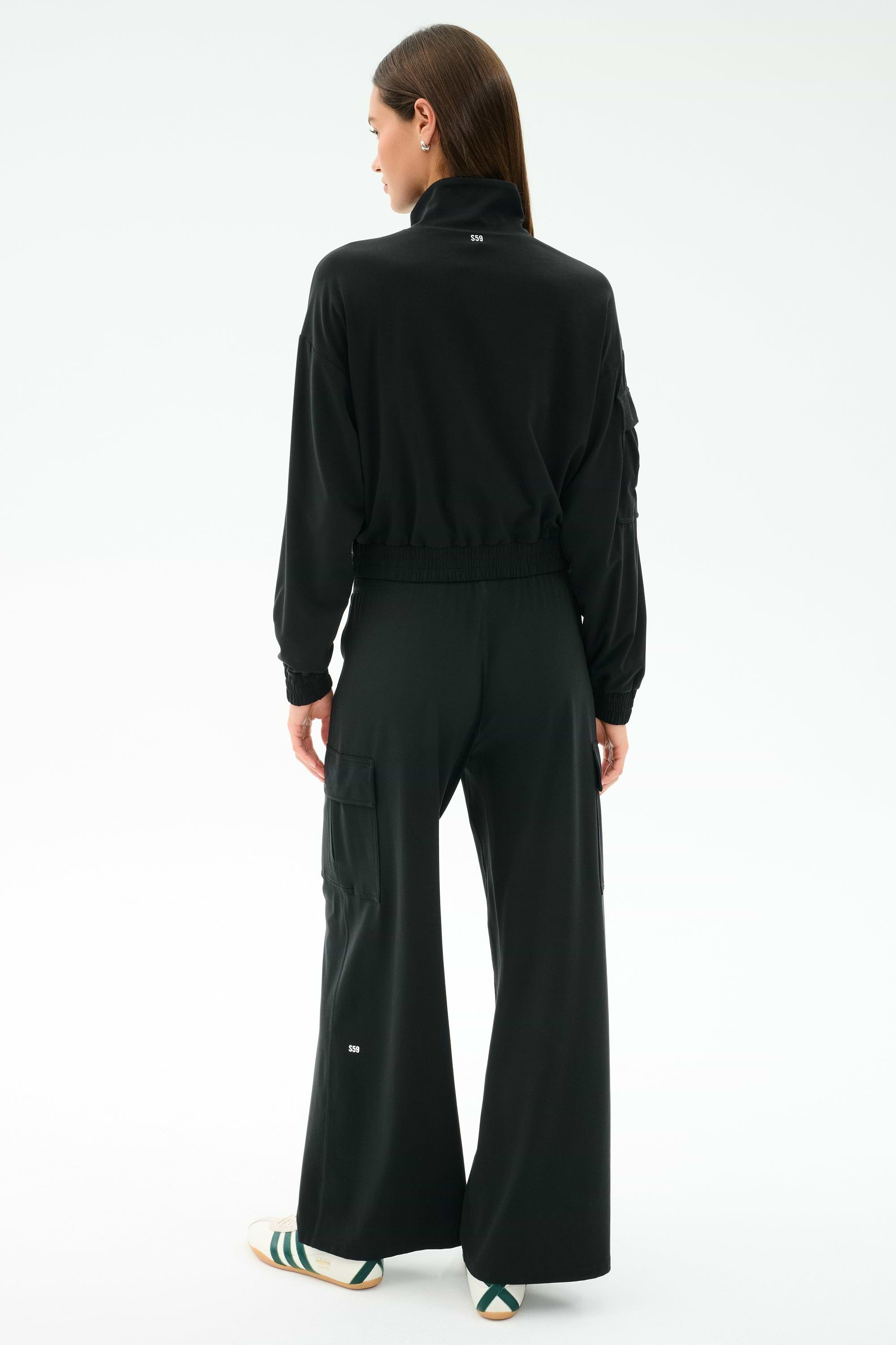 A person faces away, wearing the SPLITS59 Peaches Airweight Cargo Jacket in black stretch fabric and wide-leg pants with white text, plus white slide sandals with green stripes, against a plain light background.
