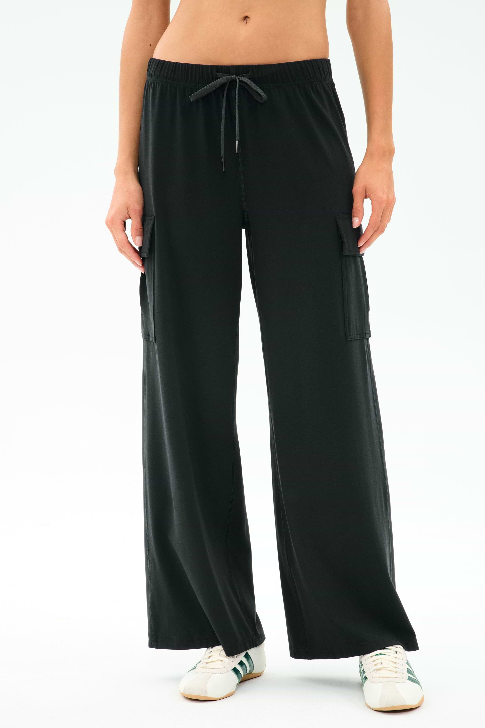 A person wearing SPLITS59’s Alex Airweight Cargo Pant in black, featuring side pockets and soft Airweight fabric, paired with white sneakers and standing against a plain light background.
