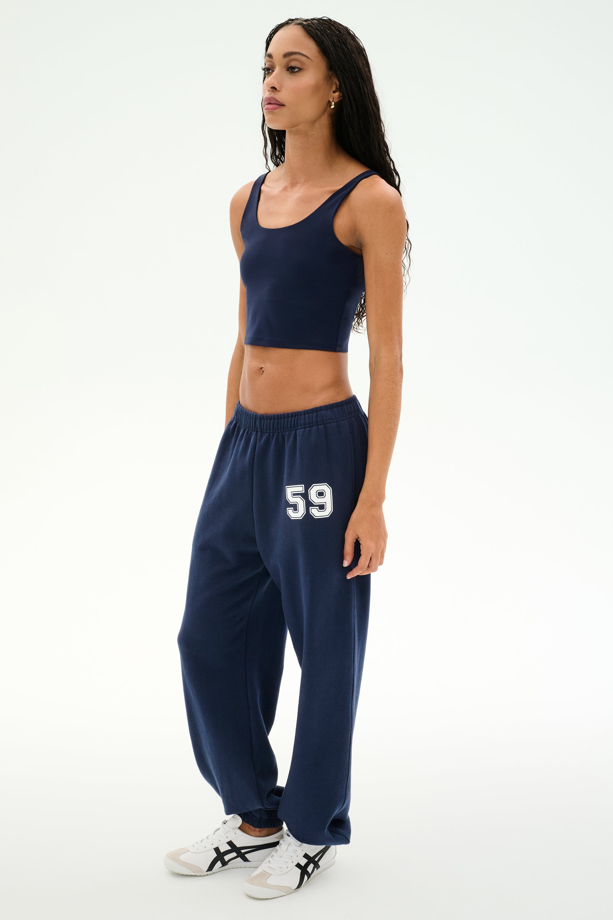 A woman is standing in SPLITS59's Serena French Terry Oversized Sweatpant in Indigo/59, crafted from soft cotton fabric, paired with a navy crop top and white sneakers against a plain white background.