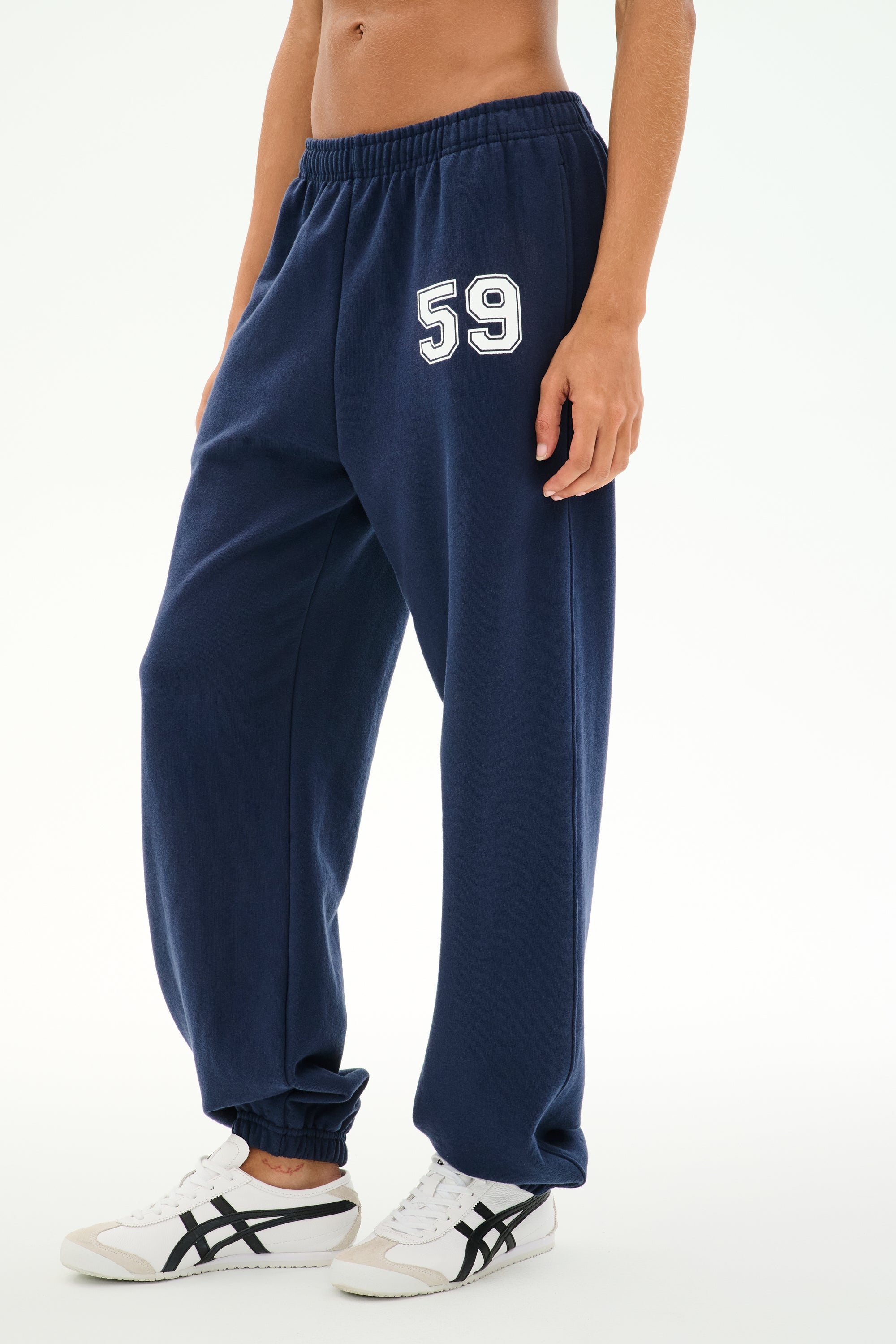A person is wearing the SPLITS59 Serena French Terry Oversized Sweatpant in Indigo/59—crafted from 100% cotton, featuring the number 59 on the left thigh—paired with white sneakers with black stripes, standing against a plain background.