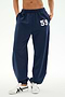 A person is seen wearing SPLITS59's Serena French Terry Oversized Sweatpant in Indigo, distinguished by the number 59 on the left thigh. The sweatpants are made from soft cotton fabric and are paired with white sneakers. Only the lower torso and legs are visible.