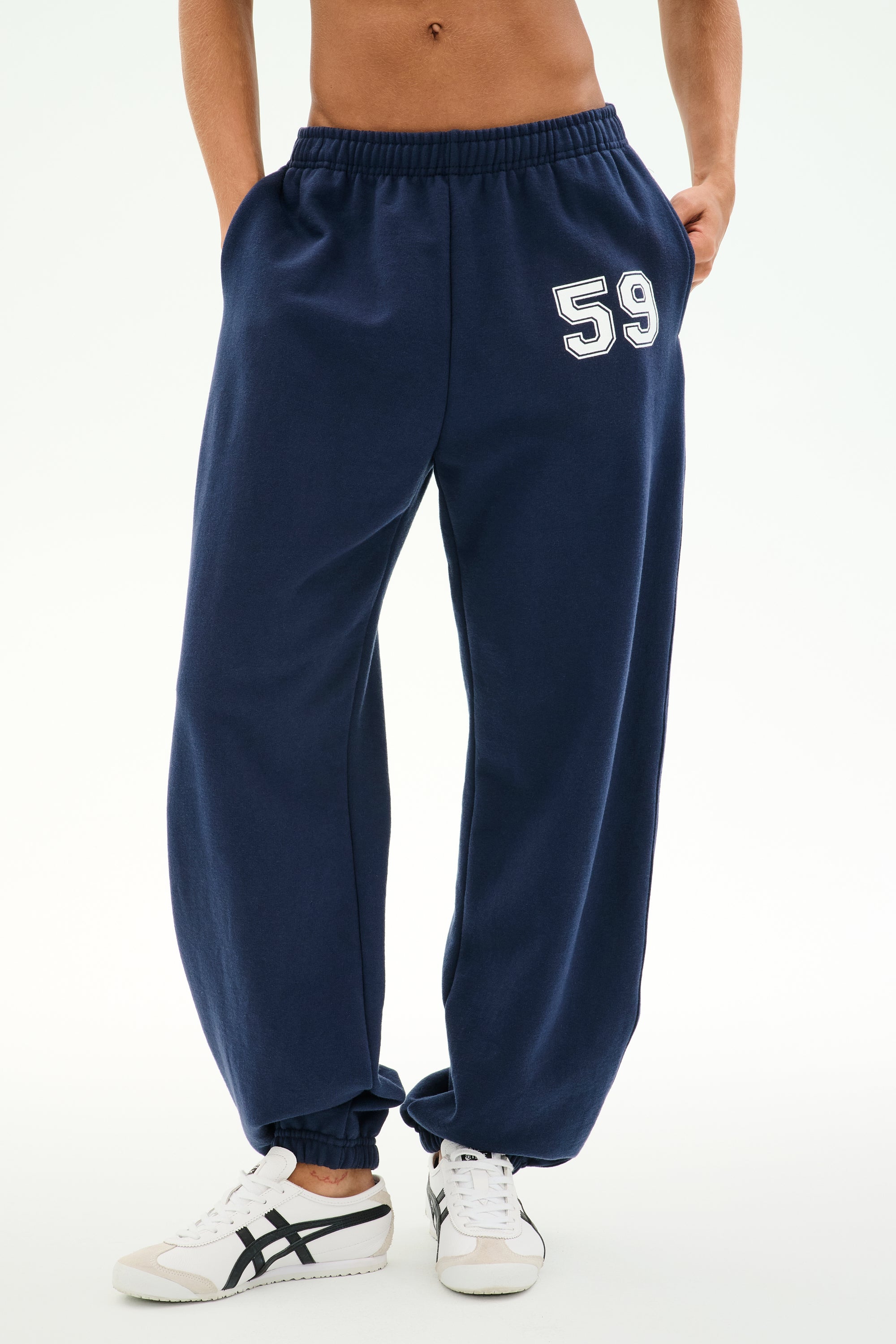 A person is seen wearing SPLITS59's Serena French Terry Oversized Sweatpant in Indigo, distinguished by the number 59 on the left thigh. The sweatpants are made from soft cotton fabric and are paired with white sneakers. Only the lower torso and legs are visible.