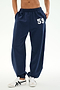 A person is seen wearing SPLITS59's Serena French Terry Oversized Sweatpant in Indigo, distinguished by the number 59 on the left thigh. The sweatpants are made from soft cotton fabric and are paired with white sneakers. Only the lower torso and legs are visible.