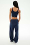 A person with long braided hair stands facing away, wearing the SPLITS59 Serena French Terry Oversized Sweatpant in Indigo/59—loose-fitting and crafted from soft cotton—paired with a navy sleeveless crop top and white sneakers against a plain light background.