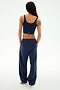 A person with long braided hair stands facing away, wearing the SPLITS59 Serena French Terry Oversized Sweatpant in Indigo/59—loose-fitting and crafted from soft cotton—paired with a navy sleeveless crop top and white sneakers against a plain light background.