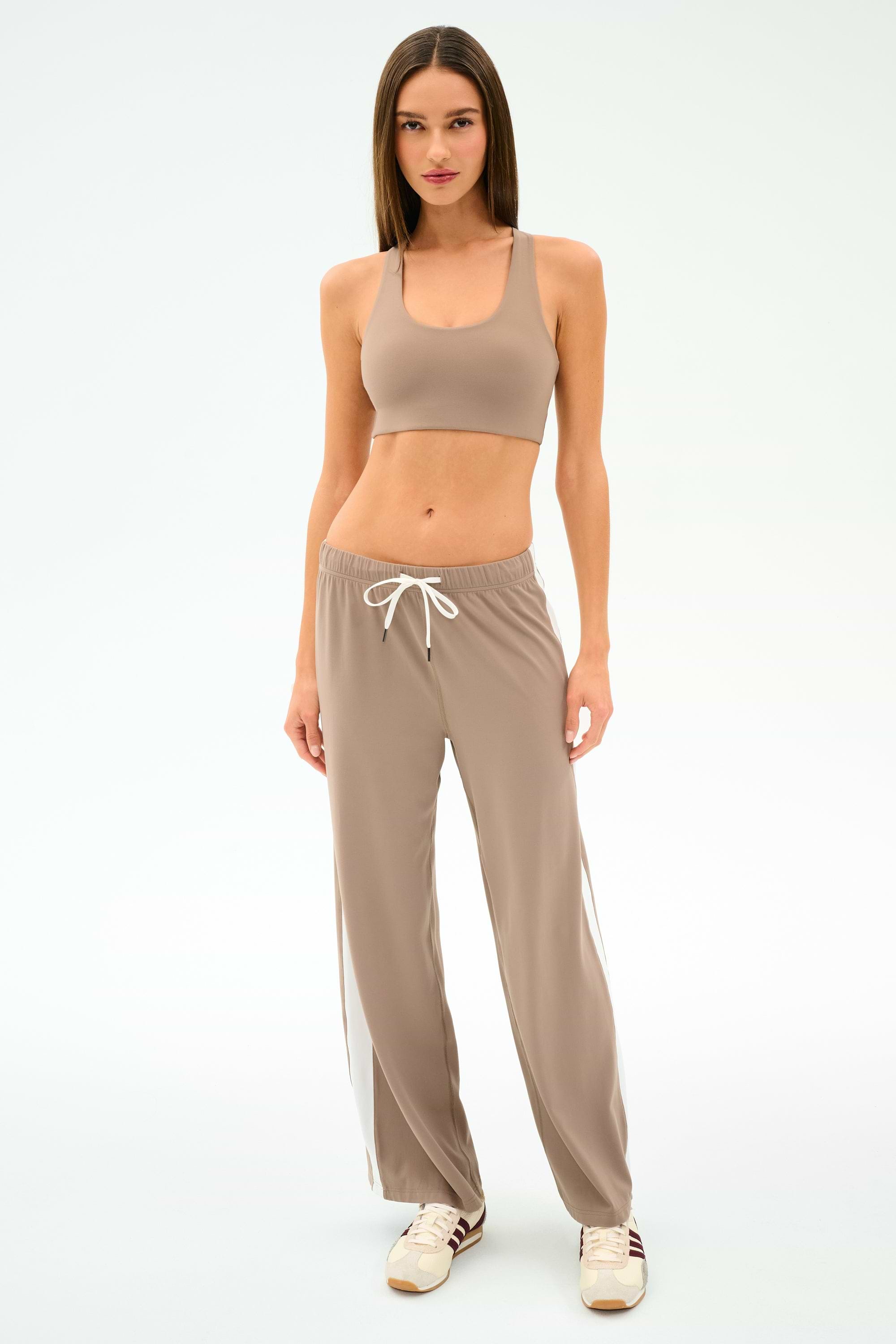 A woman faces forward in SPLITS59 Willow Airweight Wide Leg pants in Latte/White, paired with a taupe spandex sports bra and beige sneakers against a plain white background.