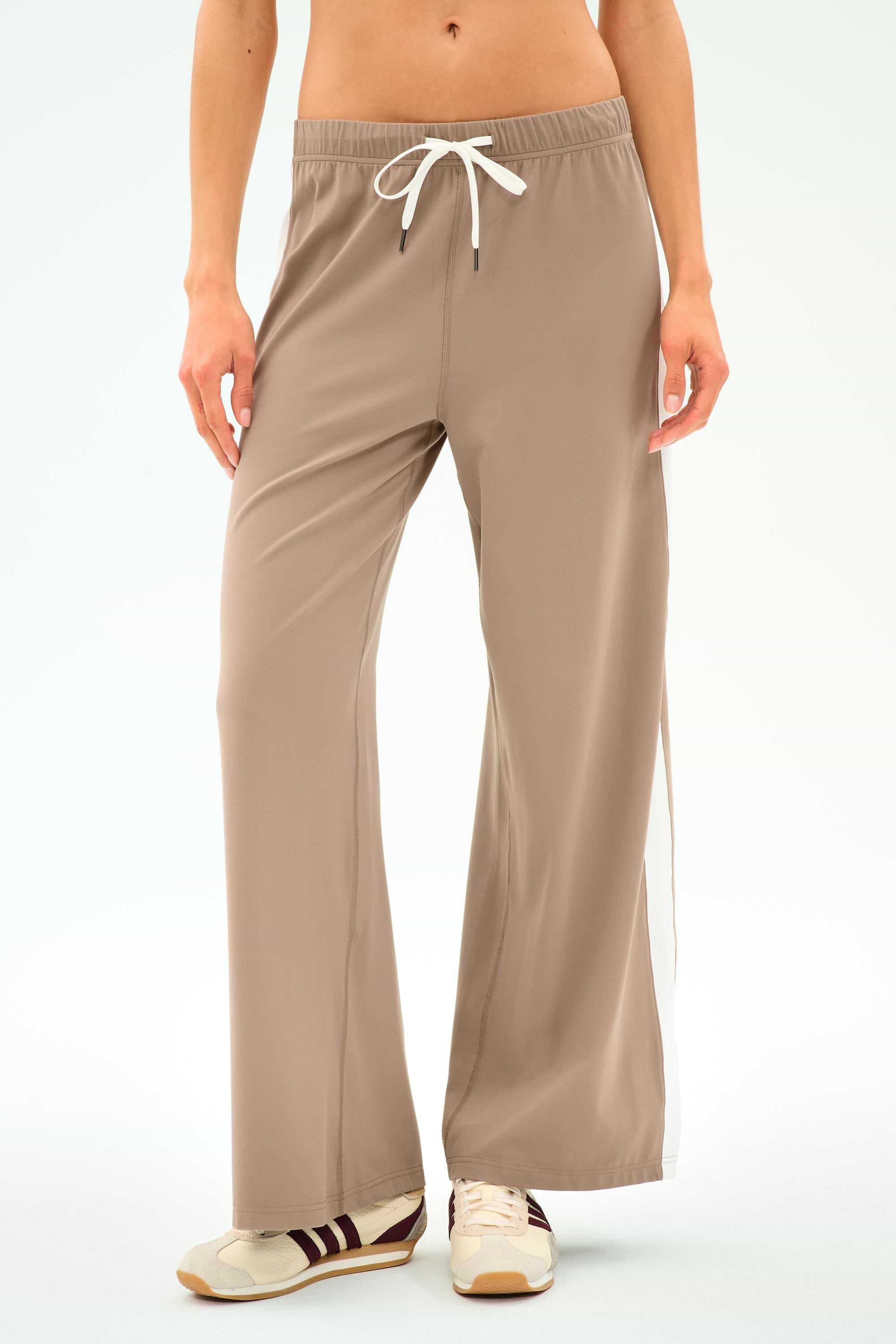 A person wears SPLITS59 Willow Airweight Wide Leg pants in Latte/White, paired with beige sneakers with brown stripes, standing against a plain white background.