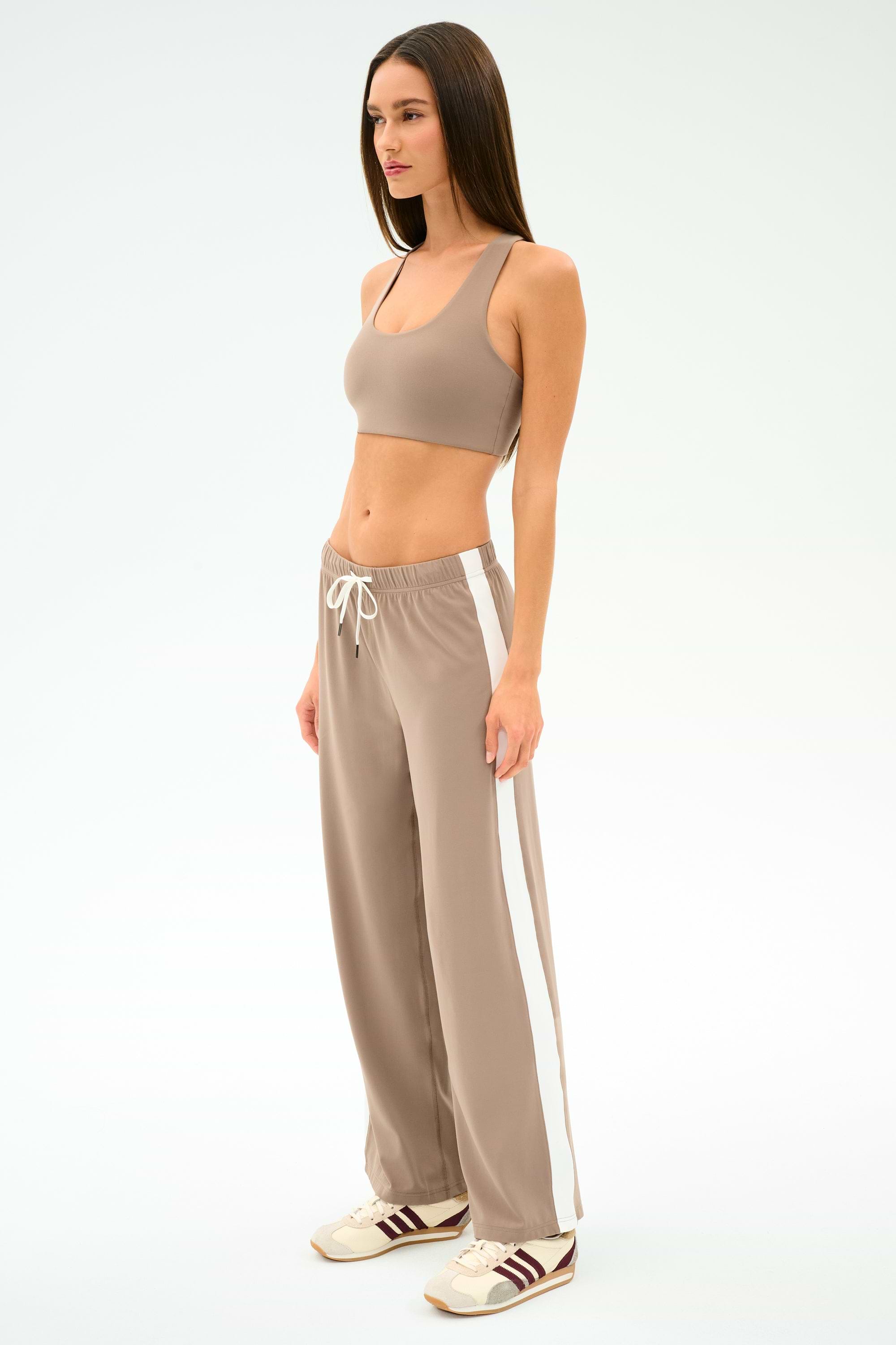 A woman stands in profile wearing SPLITS59’s Willow Airweight Wide Leg pants in Latte/White, a beige sports bra, and beige sneakers with brown stripes.